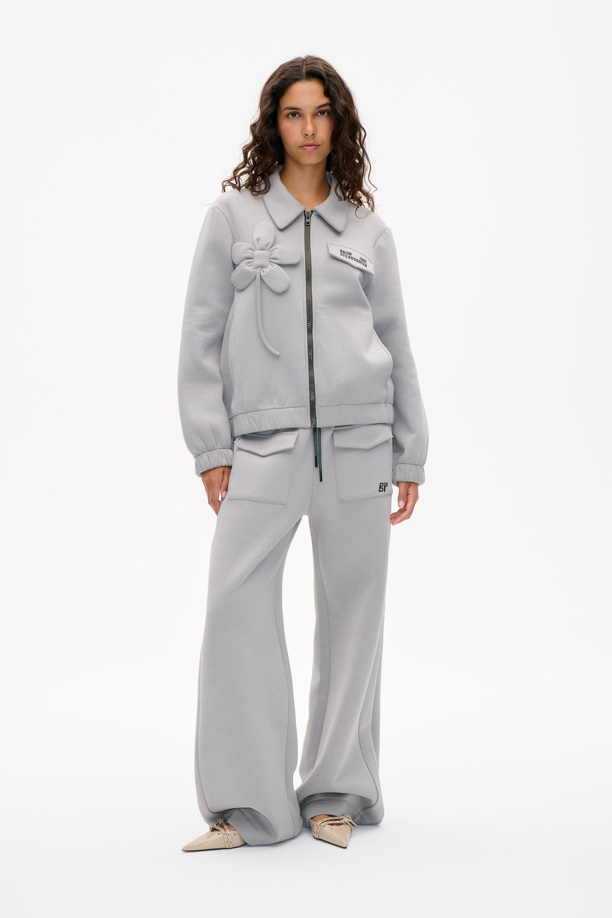 Model draagt een lichtgrijs jasje met ritssluiting vooraan, oversized kraag en bloemenapplicatie, gecombineerd met een bijpassende broek met wijde pijpen.