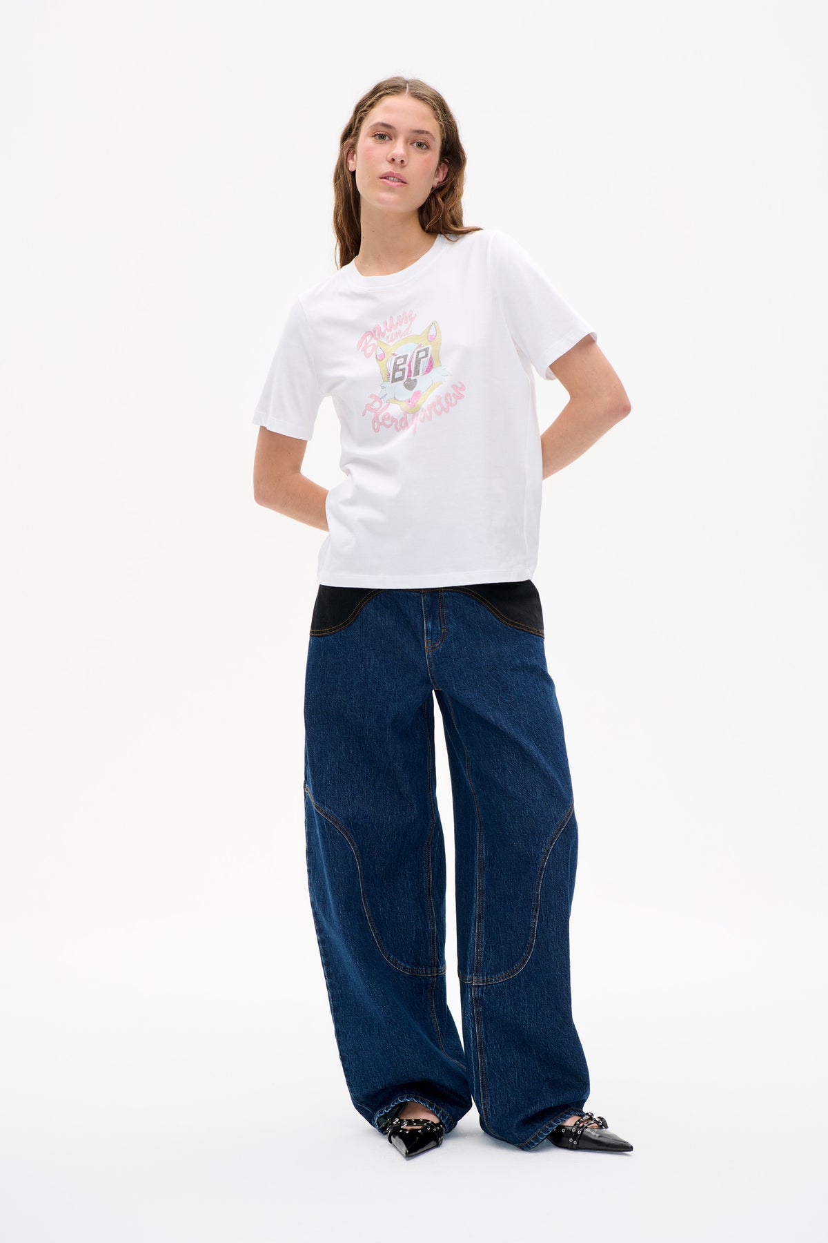 Model wearing a white short-sleeve T-shirt with a pastel cartoon and Baum und Pferdgarten lettering, styled with wide-leg dark blue jeans.