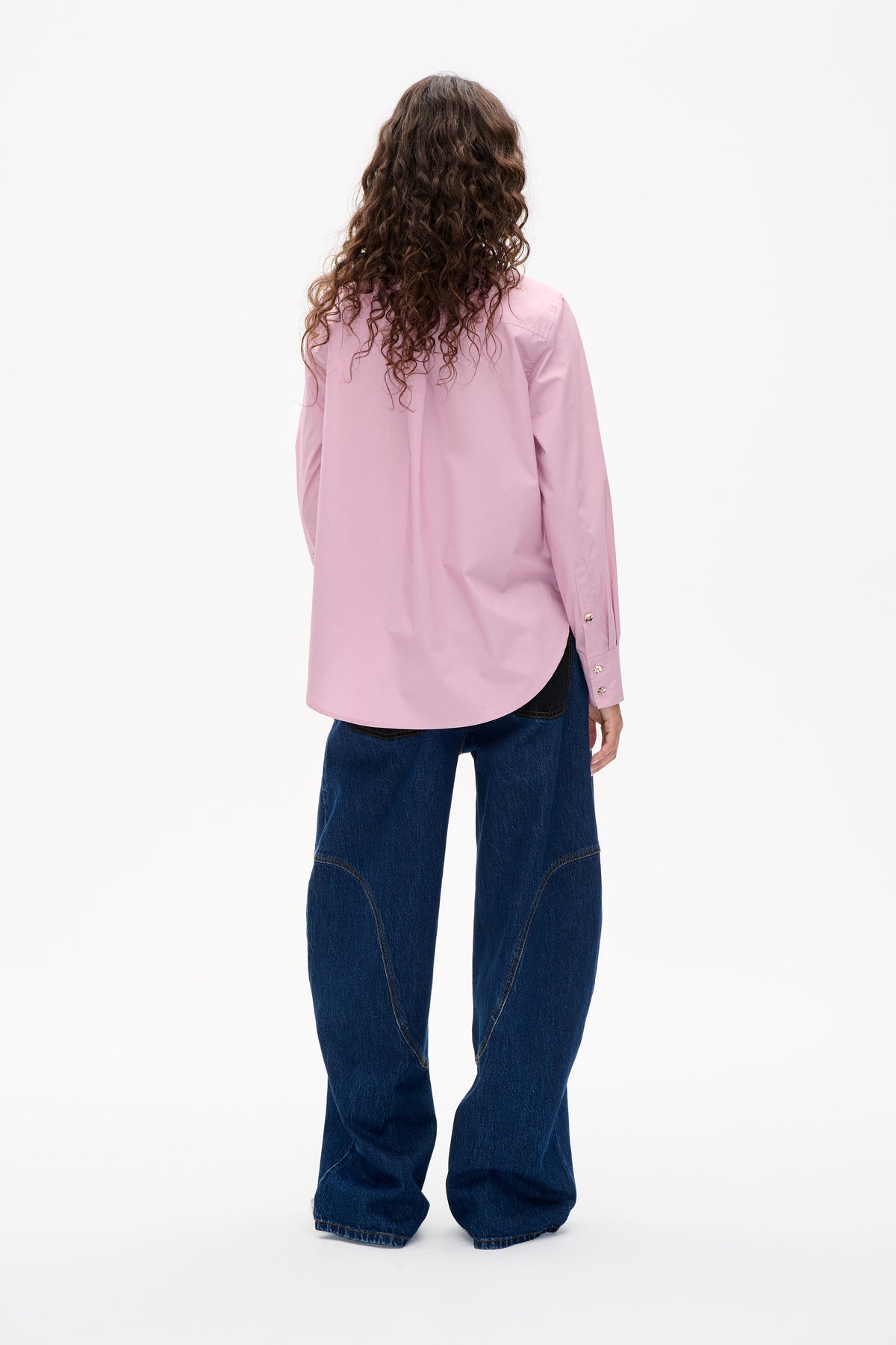 Back view of a model wearing a plain light pink long-sleeved shirt with button cuffs and dark blue wide-leg jeans.
