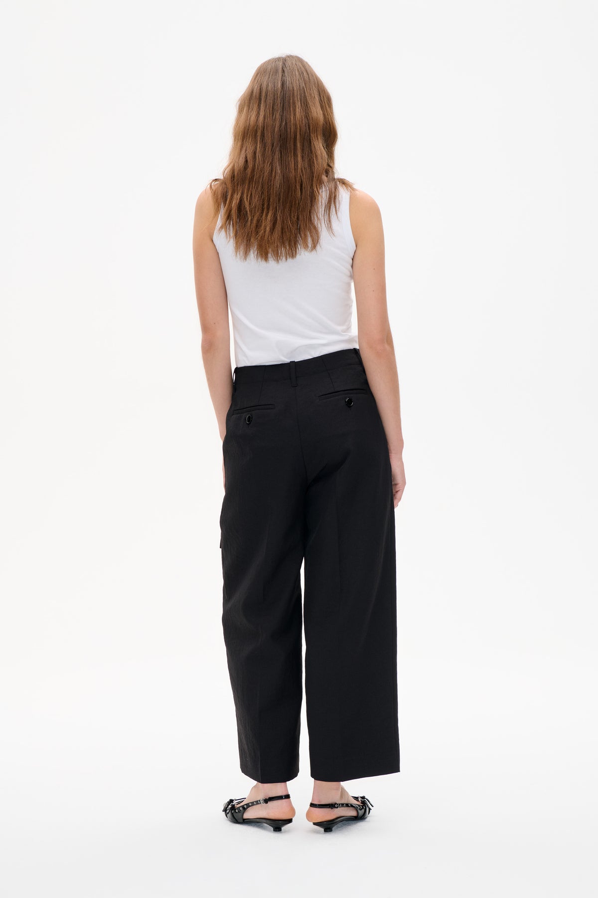 Rückansicht eines Models, das ein schlichtes weißes ärmelloses Top, eine schwarze, weitgeschnittene knöchellange Hose mit Leistentaschen und flache schwarze Riemchensandalen trägt.