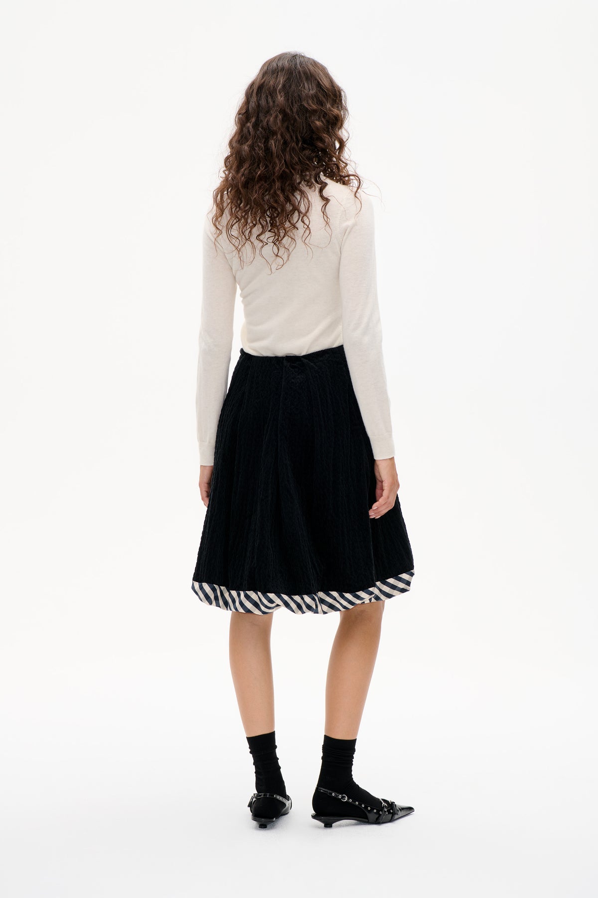 Back view of a model wearing a black textured skirt with a black-and-white gingham hem, paired with an ivory long-sleeve top.