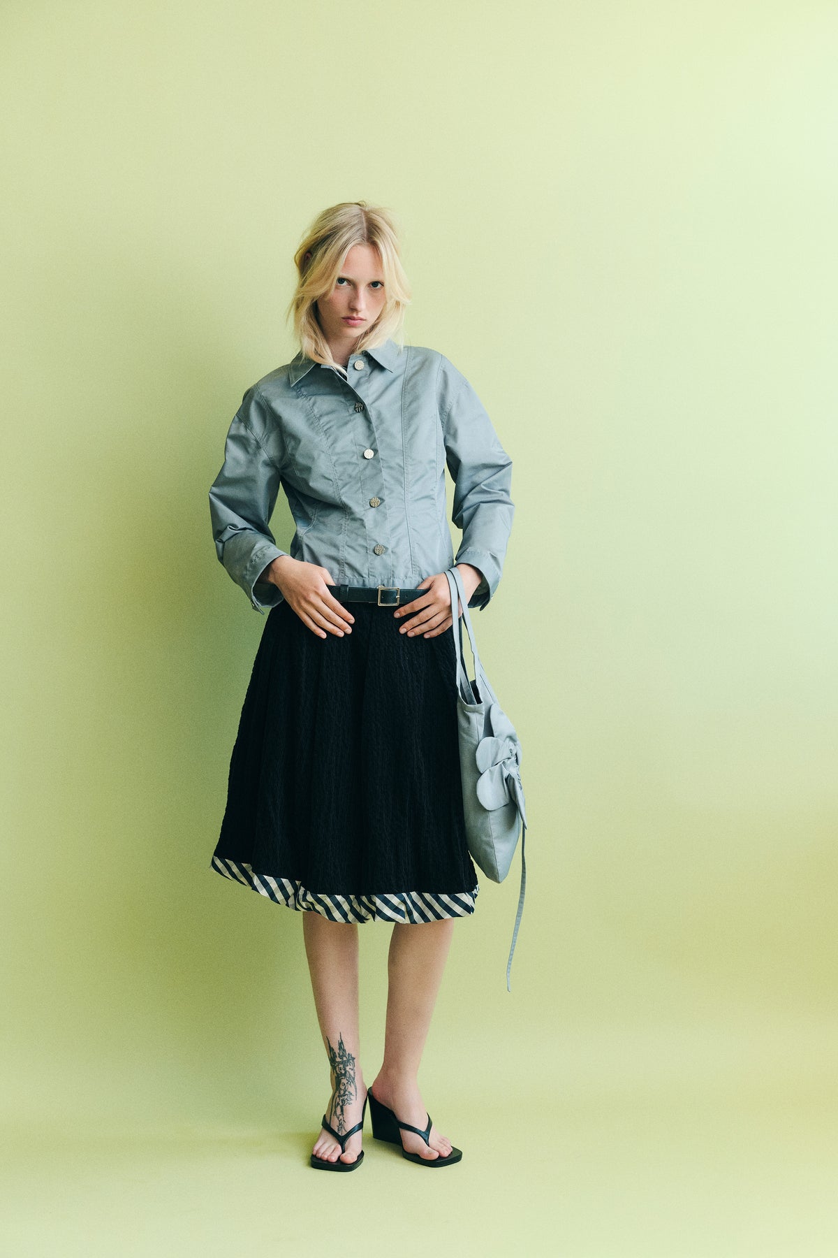 Model wearing a pale blue button-front jacket, black skirt with a checked hem and matching tote against a pale green backdrop.
