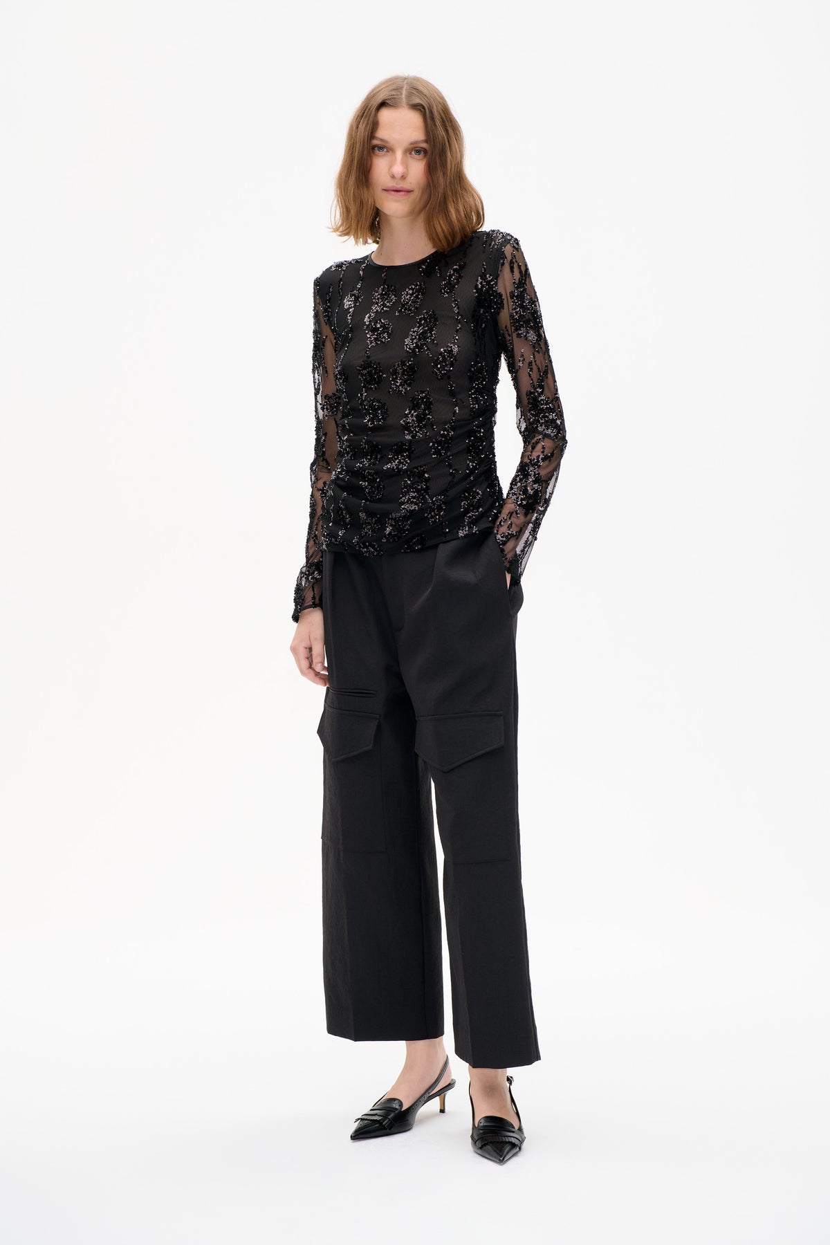 Mannequin portant un top noir transparent à manches longues, orné de motifs en sequins, et un pantalon cargo noir à jambes larges, vue de face sur fond blanc.