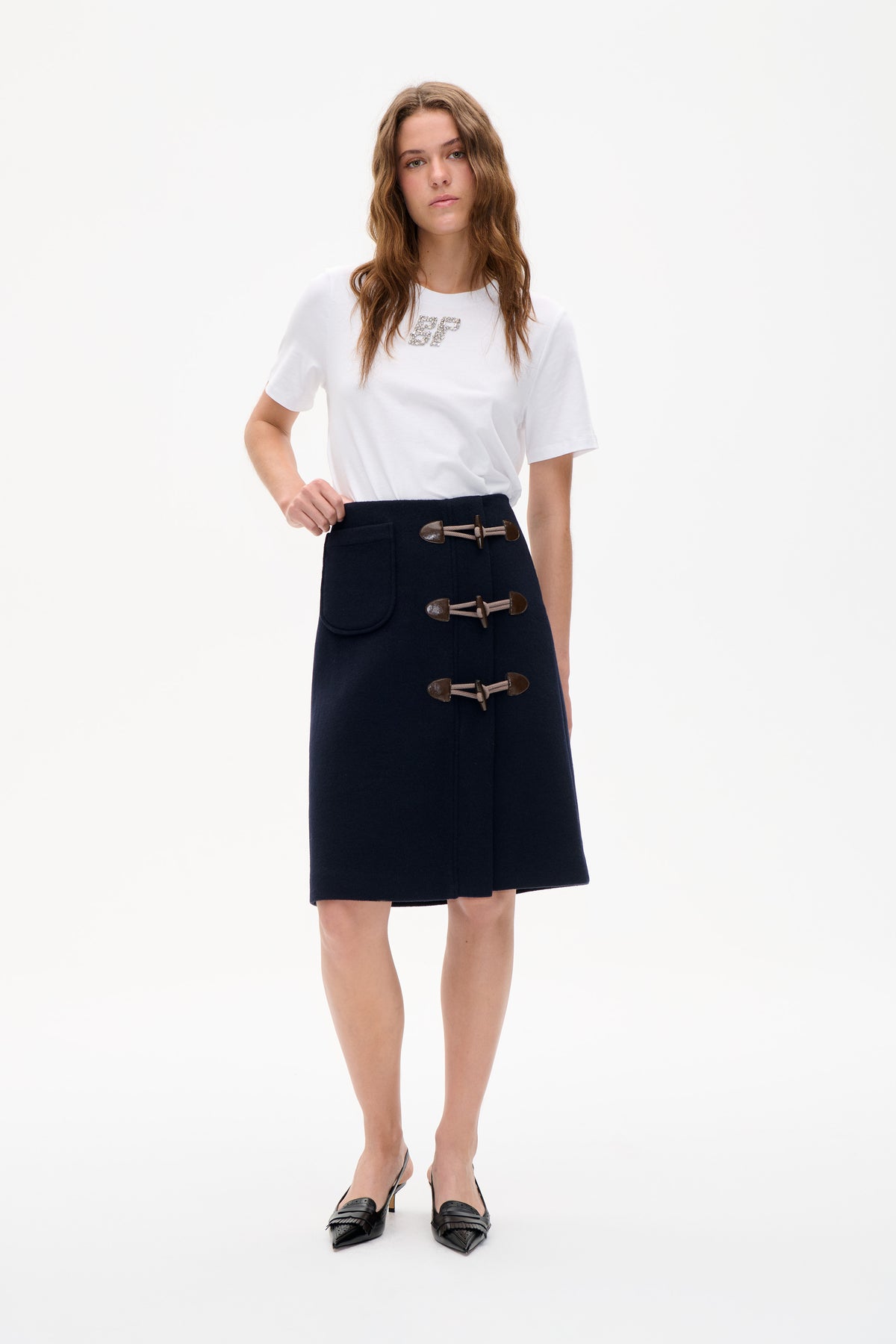 Model wearing a white short-sleeve T-shirt with a sparkling chest monogram and a navy toggle-front skirt with a patch pocket.