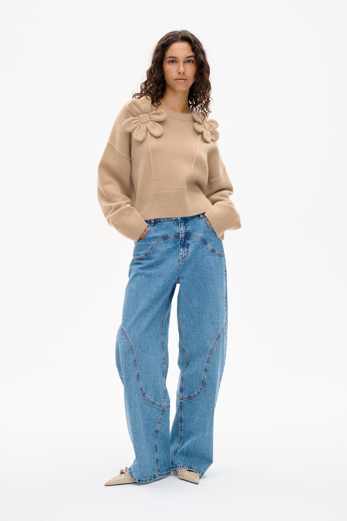 Front view of a beige knit sweater with oversized floral appliqués at the shoulders, styled with wide-leg blue jeans.