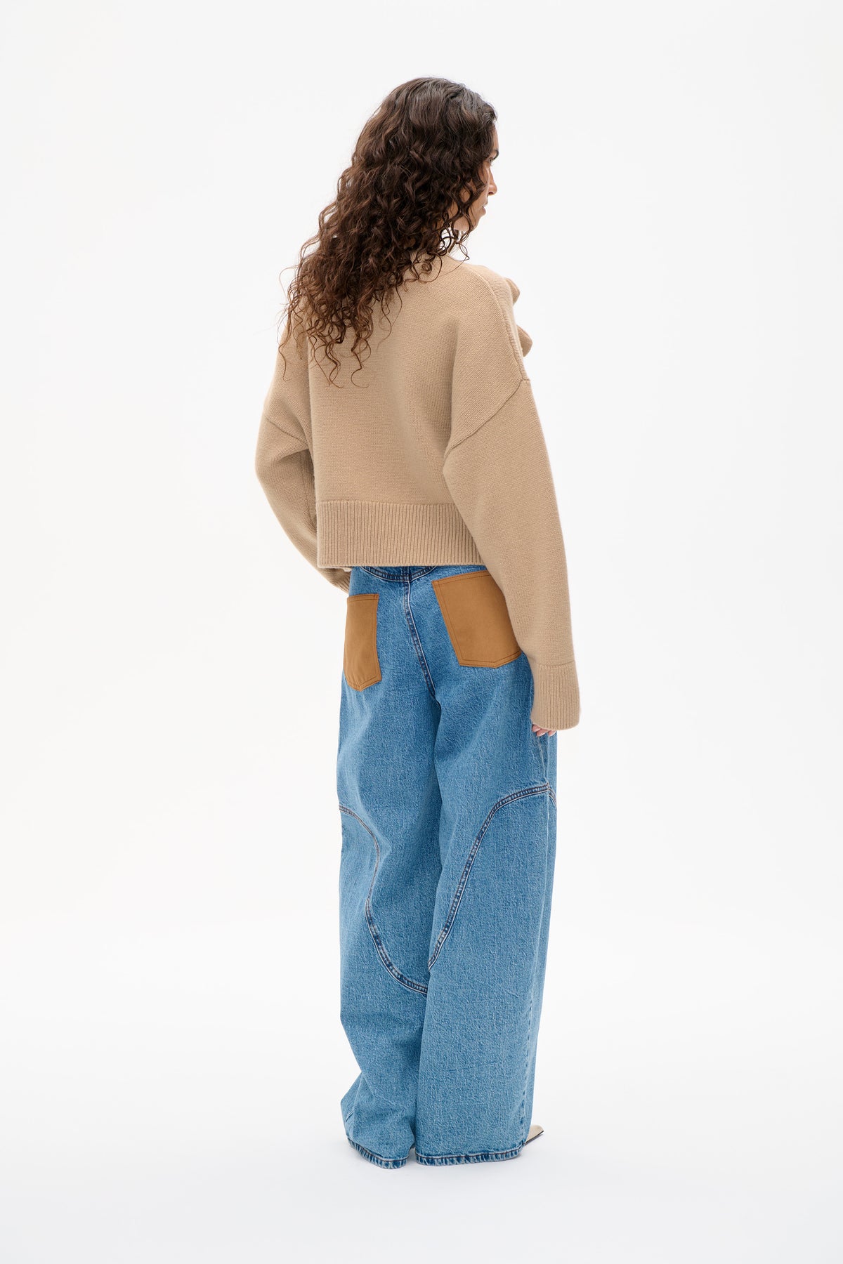 Model turned to show the back of a beige knit sweater with dropped shoulders and a ribbed hem, paired with blue jeans with tan patch pockets.