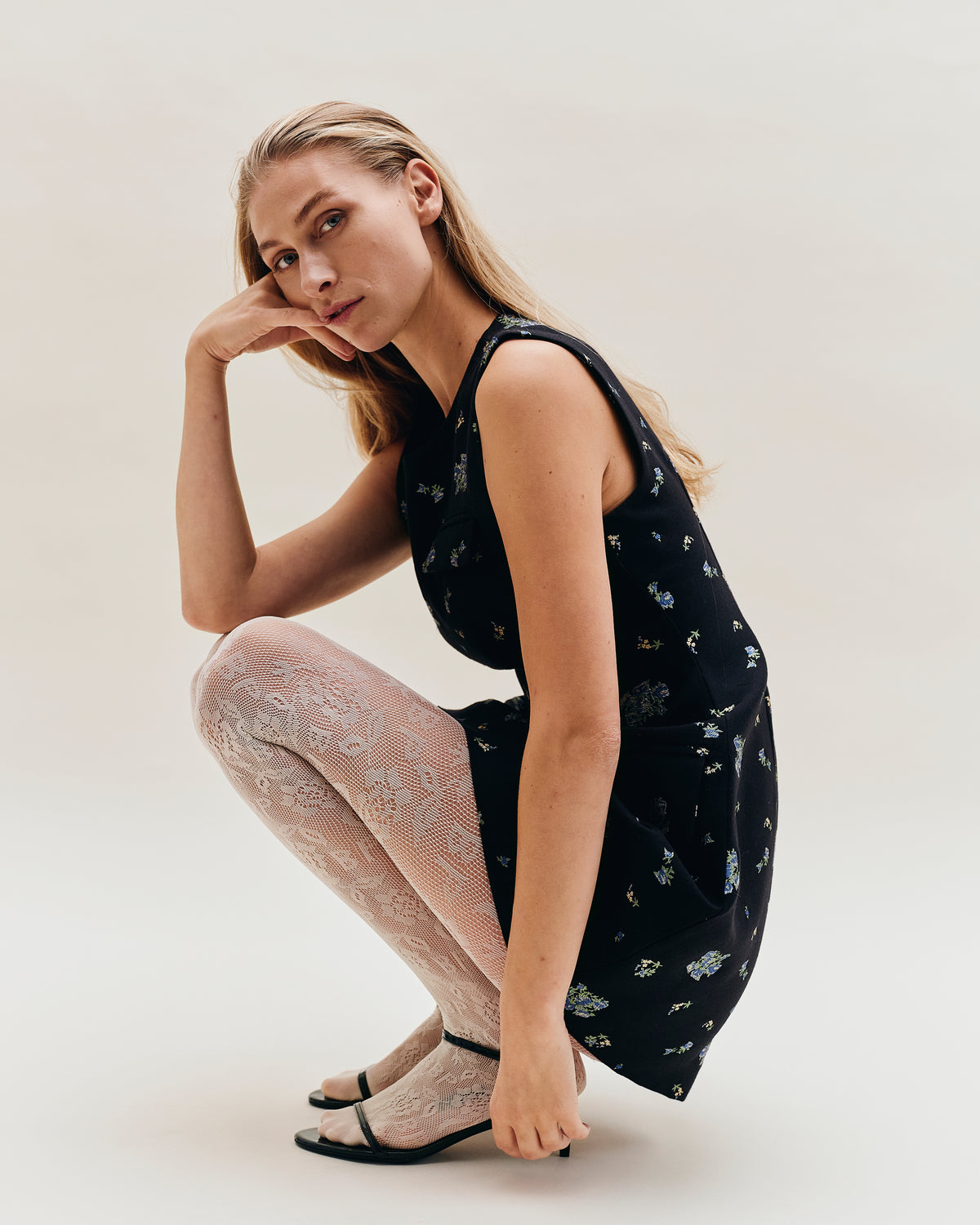 Campaign portrait of a seated model wearing a black sleeveless dress with small blue and cream flowers, pale open-knit tights and black heels.