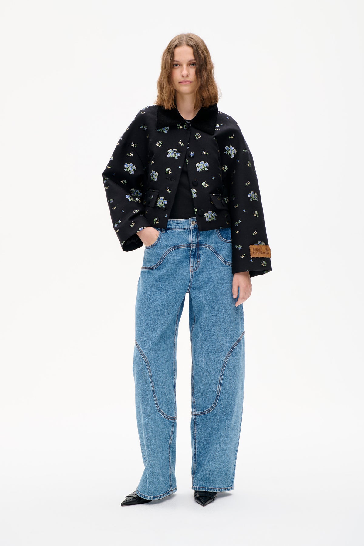 Front view of a model wearing a black jacket with scattered blue and green floral print, oversized collar and wide-leg blue jeans.