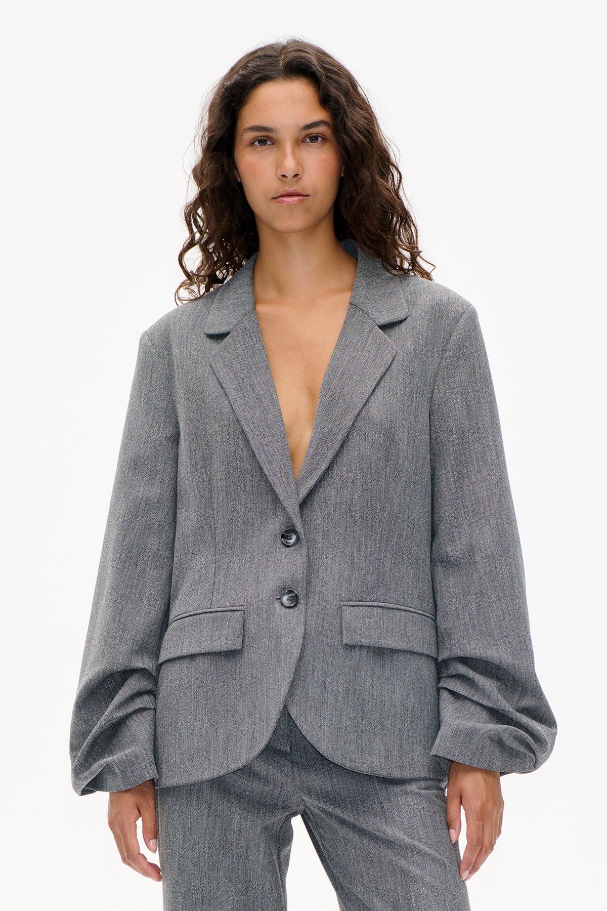 Front view of a model wearing a textured gray blazer with notched lapels, two buttons and flap pockets, paired with matching trousers.