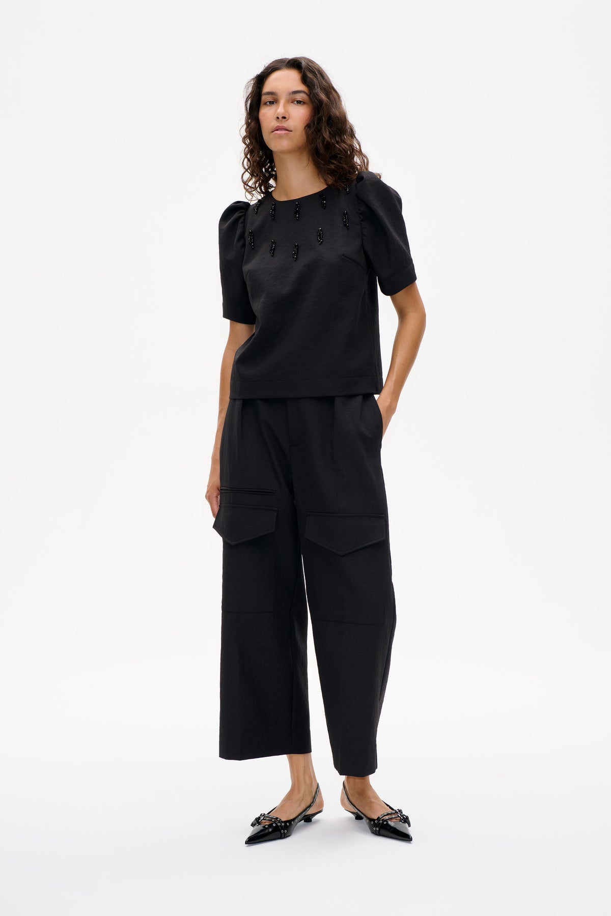 Mannequin portant un top noir à manches bouffantes et détails perlés sur le devant, associé à un pantalon raccourci à coupe large, doté de grandes poches plaquées.