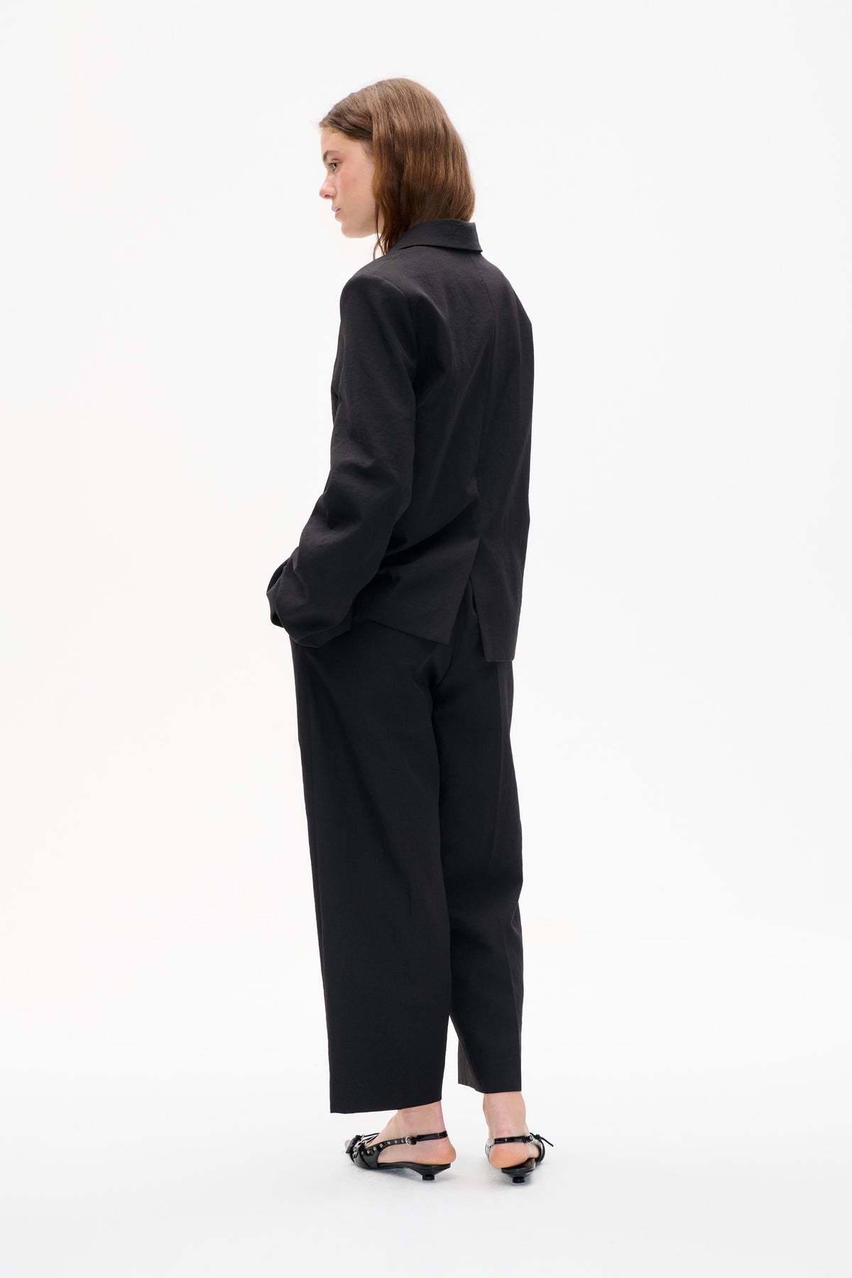 Model wearing a black collared jacket and wide-leg cropped trousers, shown from the side and back against a white studio background.
