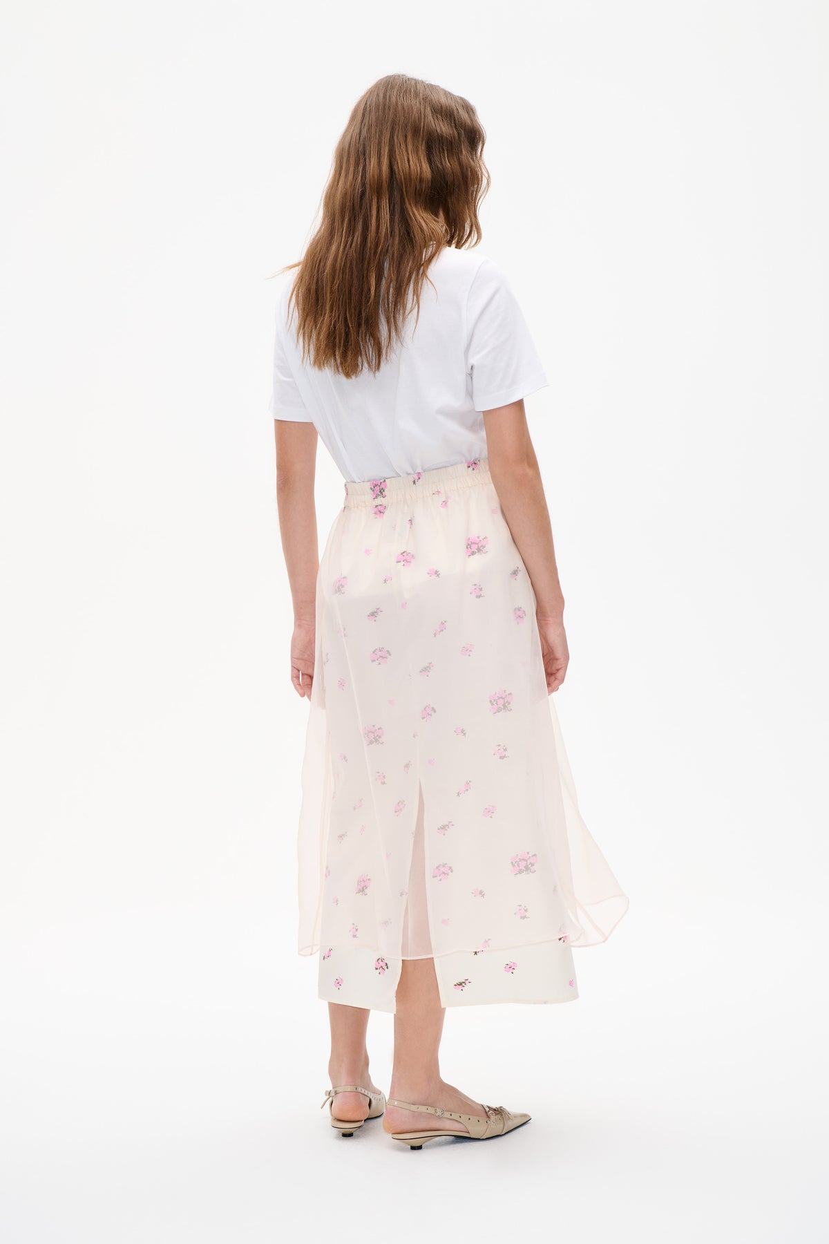 Achteraanzicht van een model in een wit T-shirt met korte mouwen en een ivoren midirok met roze bloemenprint en doorschijnende, gelaagde panelen.