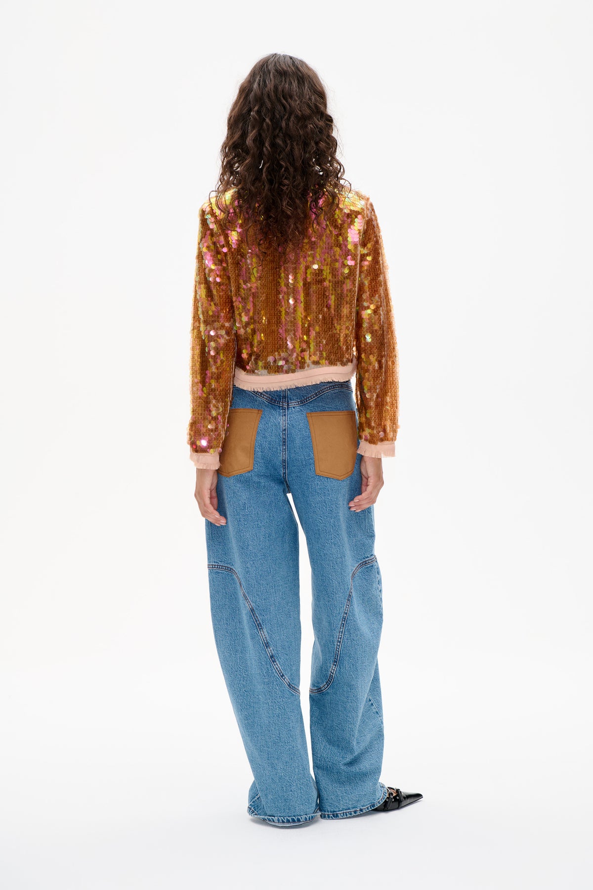 Back view of a model wearing a multicoloured sequin long-sleeved top with pale pink trim and wide-leg blue jeans with brown patch pockets.