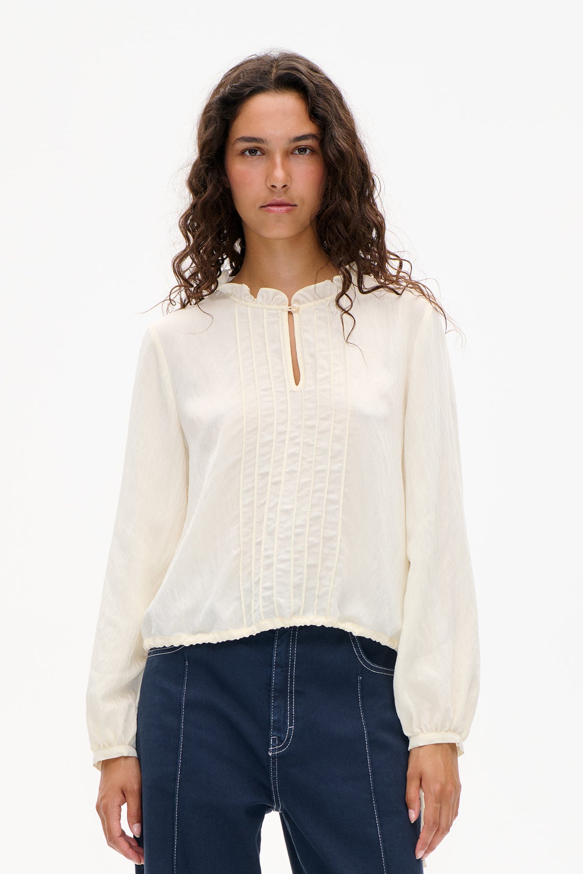 Front view of an ivory long-sleeved blouse with a ruffled collar, pintuck panel and small keyhole opening, worn with dark indigo jeans.
