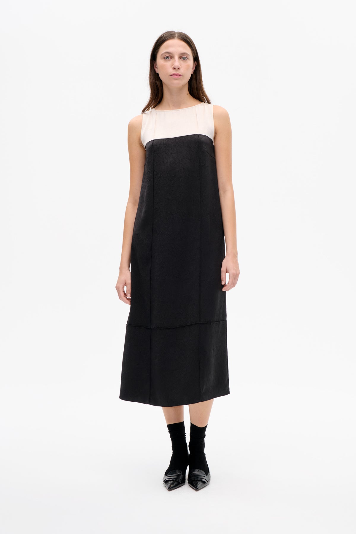 Front view of a sleeveless black midi dress with a pale blush upper panel and crinkled texture, styled with black socks and loafers.