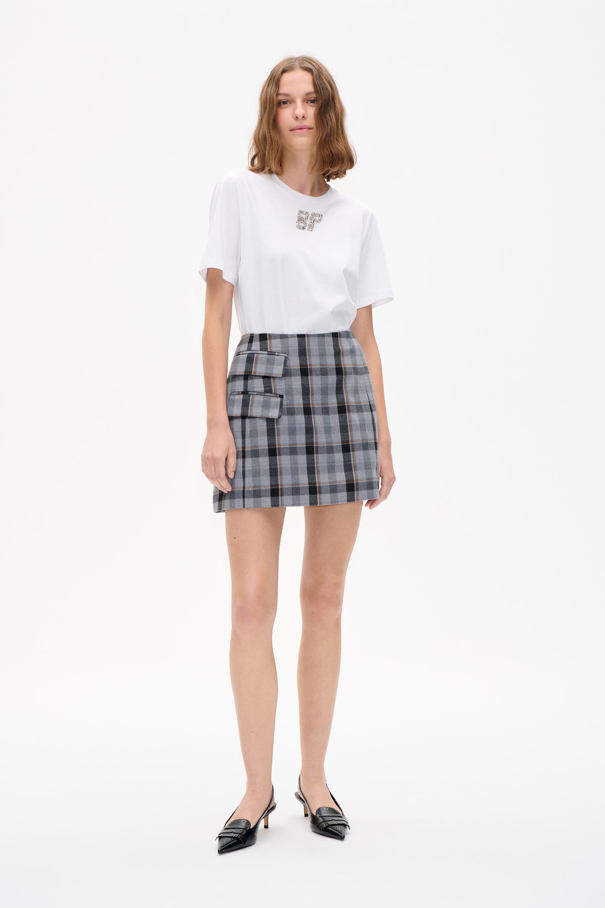 Front view of a model in a white short-sleeved T-shirt with a jeweled BP emblem and a grey plaid mini skirt with flap pockets.