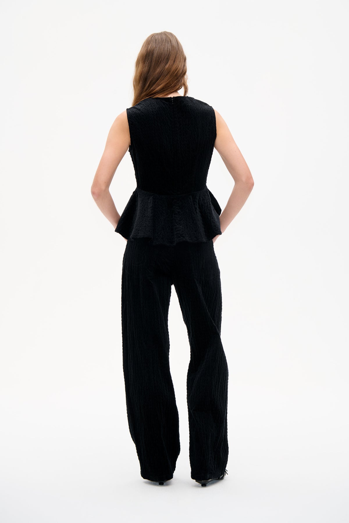 Back view of a model wearing a black sleeveless textured peplum top with a rear zipper and matching wide-leg trousers.