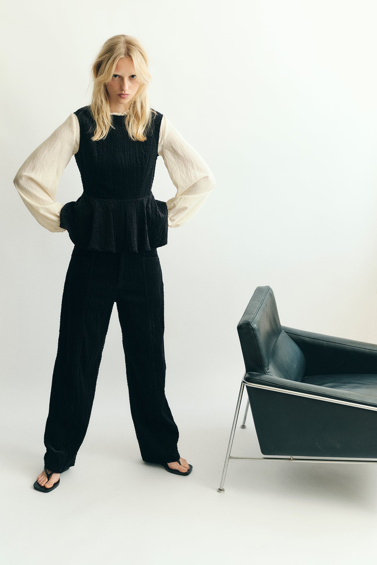 Model wears a black textured sleeveless peplum top over an ivory blouse with voluminous sleeves and matching trousers beside a black chair.