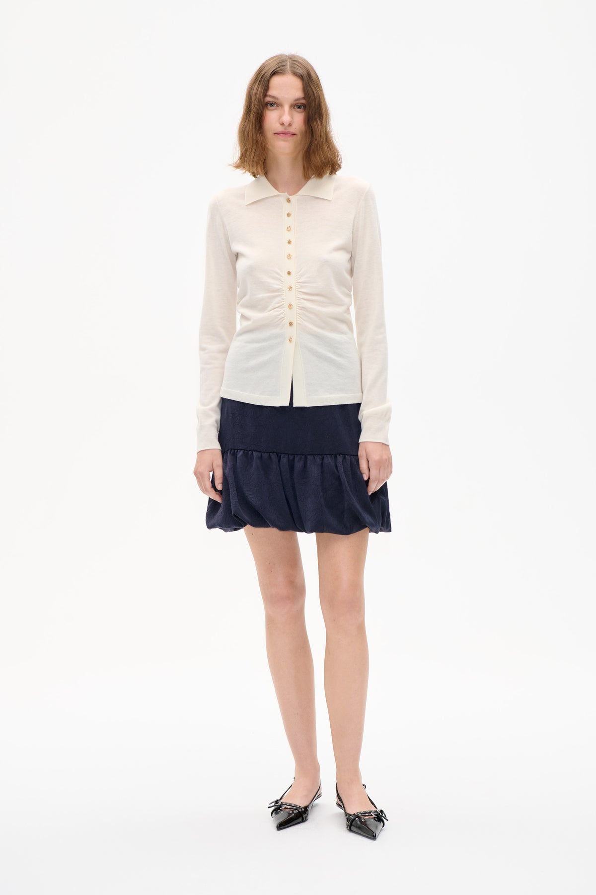 Front view of a model wearing an ivory long-sleeved knit cardigan with a pointed collar and gold decorative buttons, paired with a navy textured skirt.