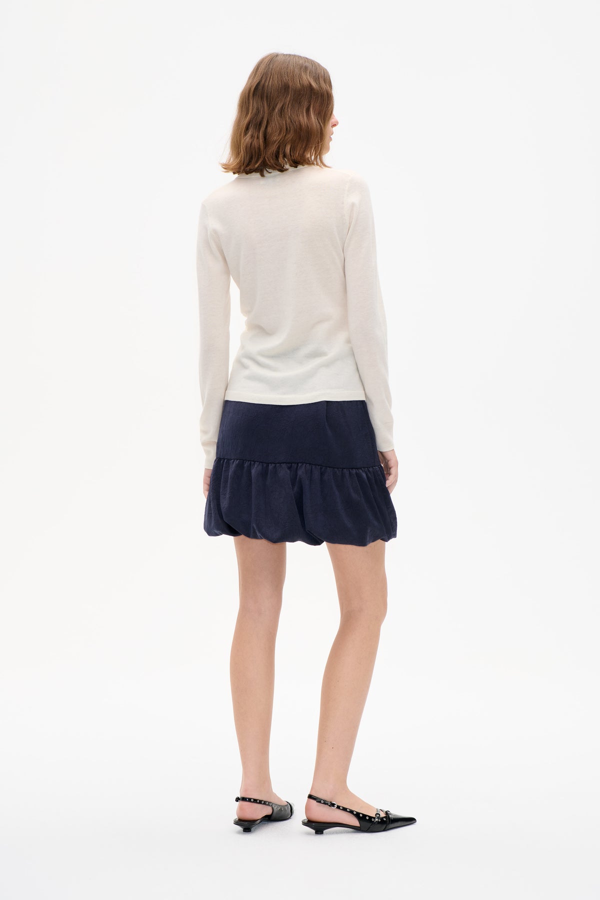 Back view of a model wearing a cream long-sleeved knit top and a navy textured mini skirt with a gathered hem.