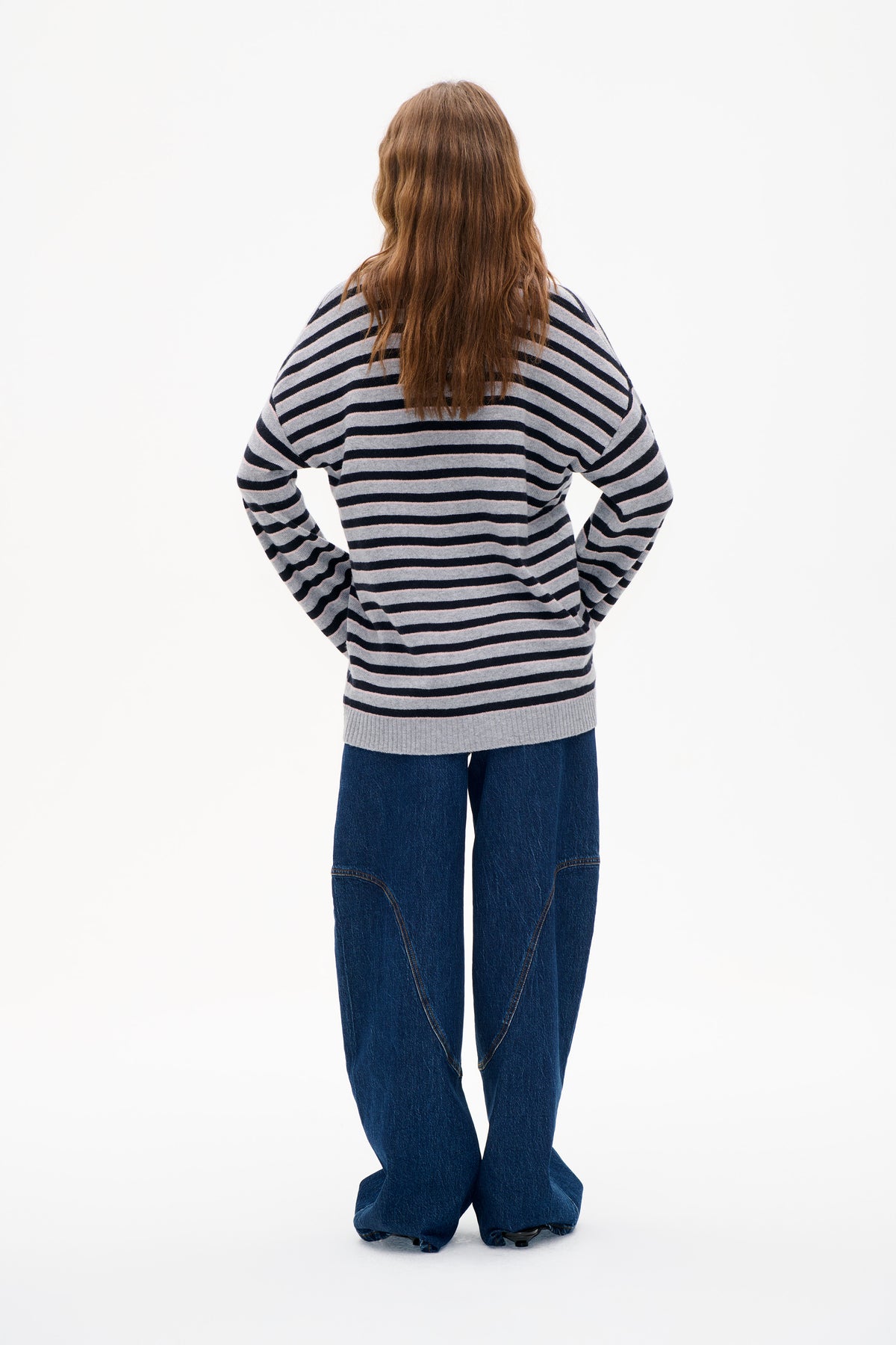 Rückenansicht eines Models, das einen grauen Strickpullover mit marineblauen und blassrosa horizontalen Streifen trägt, kombiniert mit weiten dunkelblauen Jeans.