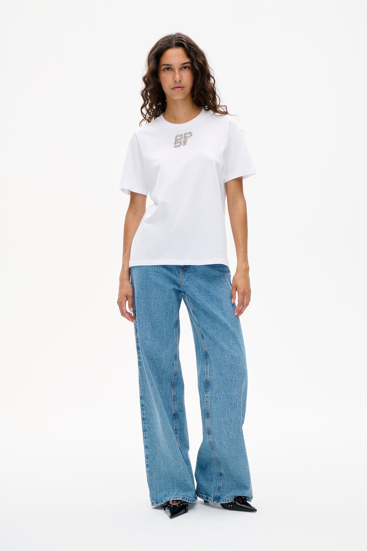 Vooraanzicht van een model in een wit T-shirt met korte mouwen en een letterembleem met sierstenen, blauwe jeans met wijde pijpen en zwarte puntige schoenen.