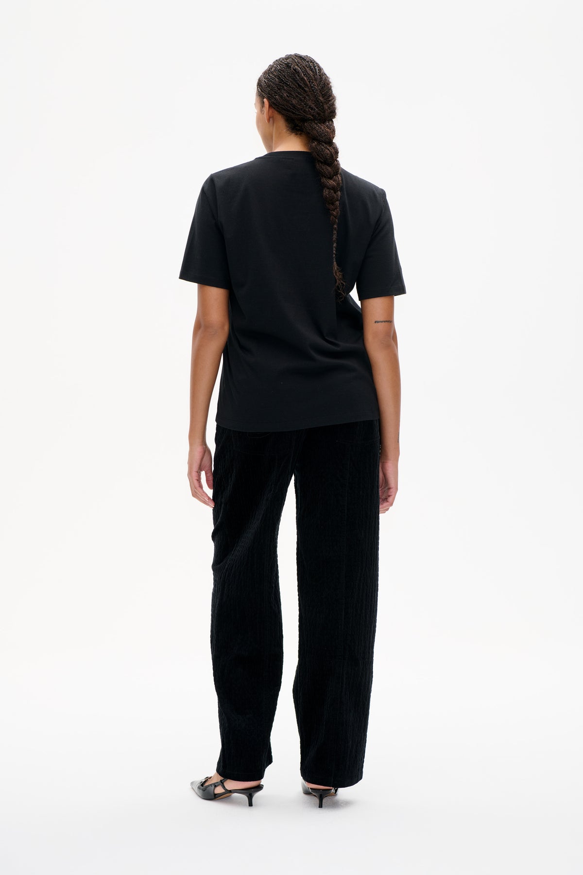 Back view of a model wearing a plain black short-sleeved T-shirt and black textured cropped trousers.