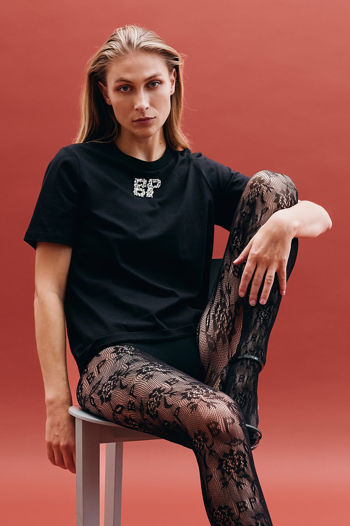 Model seated against a coral backdrop, wearing a black short-sleeve top with a sparkling chest motif and black lace-pattern tights.