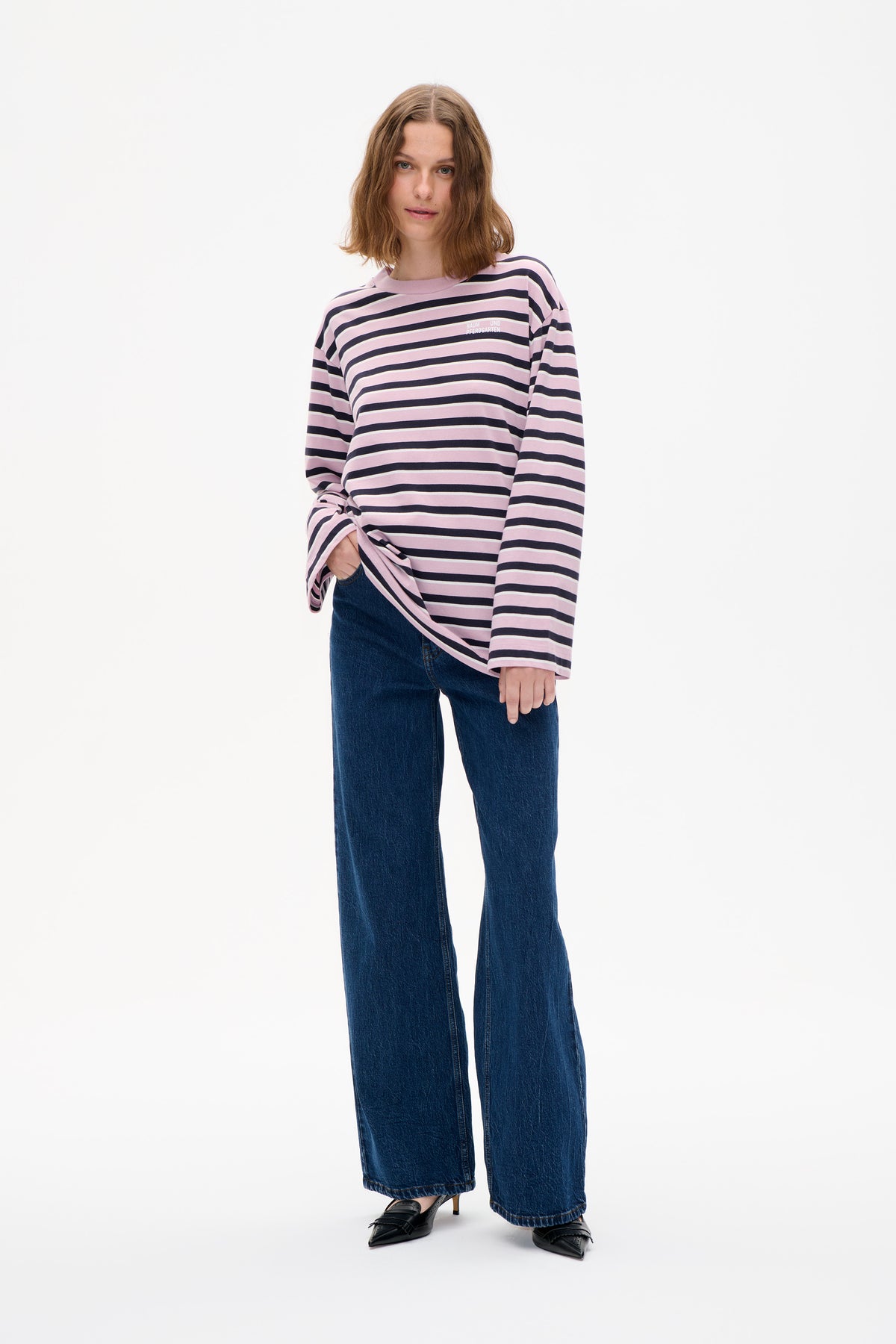 Model wearing a pink long-sleeved top with navy and cream horizontal stripes and a small chest logo, paired with wide-leg blue jeans.
