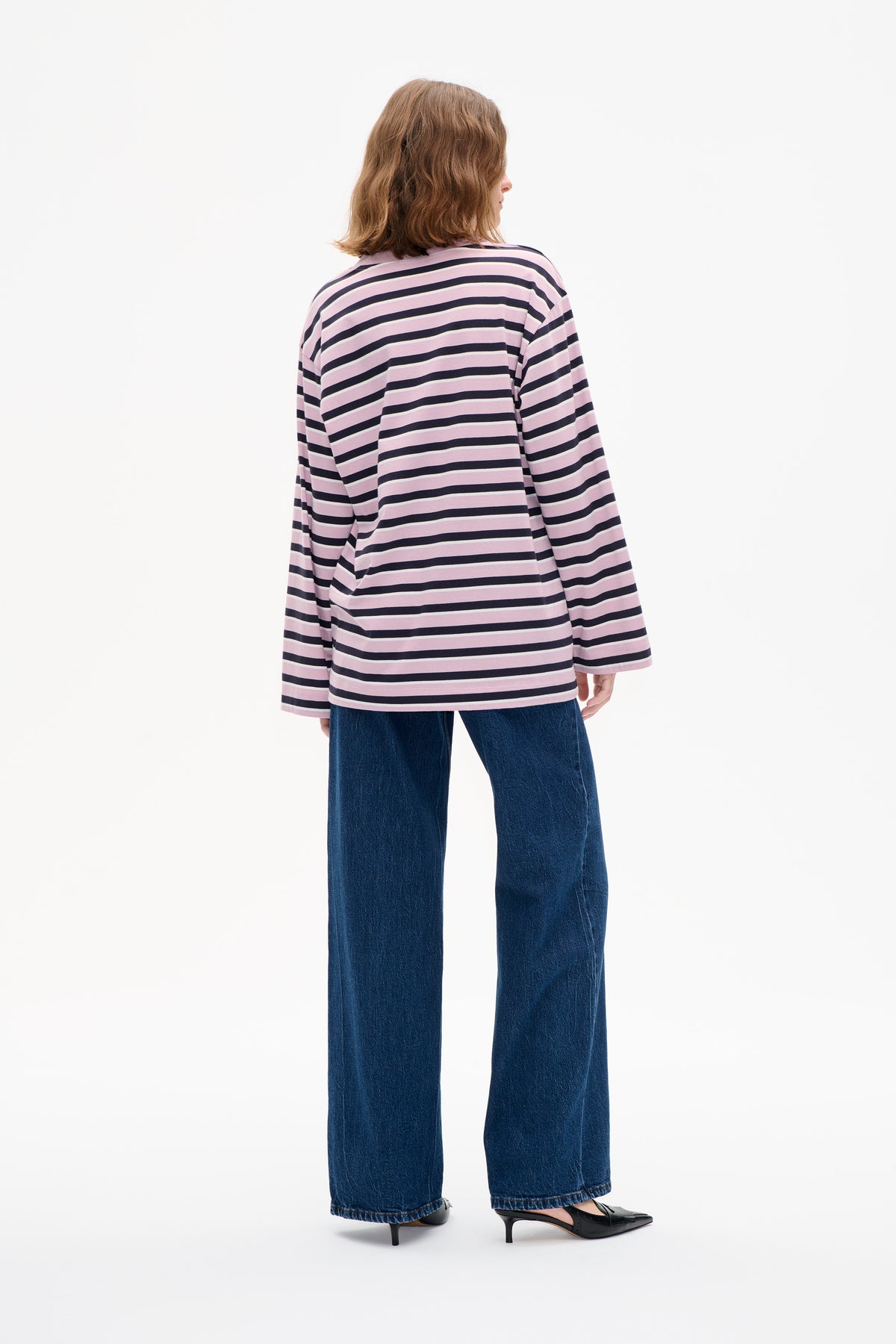 Back view of a model wearing a loose pink long-sleeved top with navy and thin cream horizontal stripes, paired with wide-leg blue jeans.