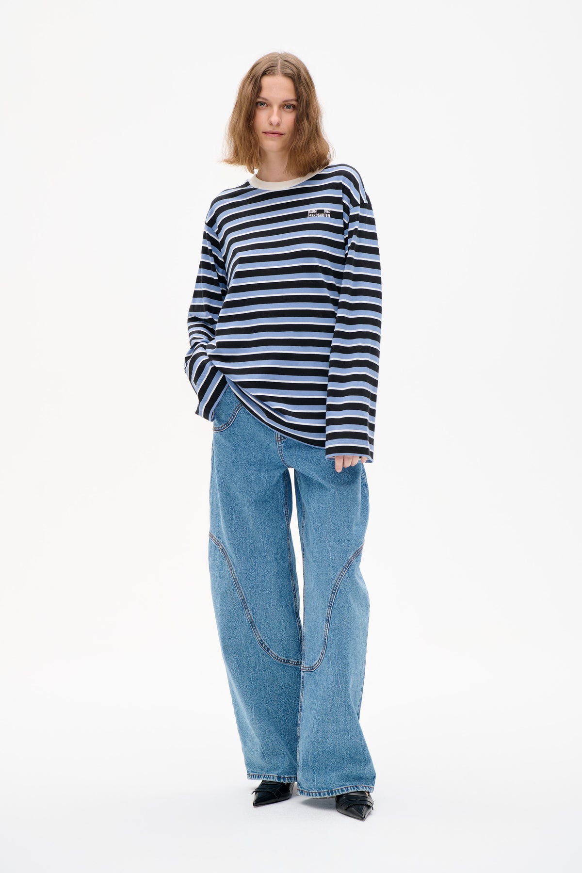 Vooraanzicht van een model dat een gestreepte top in zwart, lichtblauw en wit draagt met lange mouwen, een klein logo op de borst en wijd uitlopende blauwe jeans.