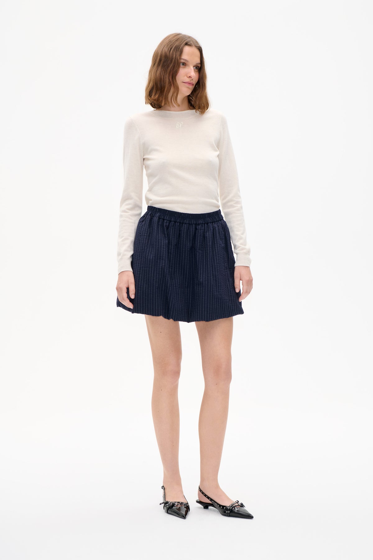 Model wearing an ivory long-sleeve crew-neck top with a small chest emblem and navy pinstripe shorts against a white studio background.