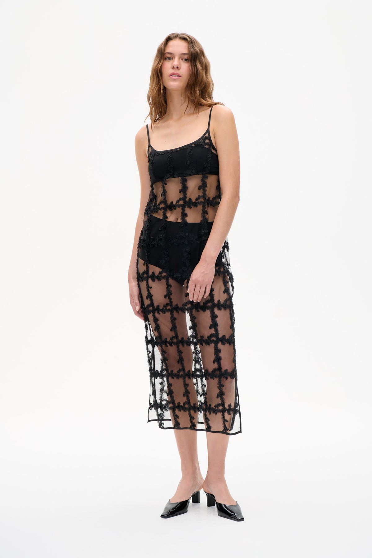 Model wearing a black sheer slip dress with thin straps, an opaque bodice and raised floral lace pattern against a white background.
