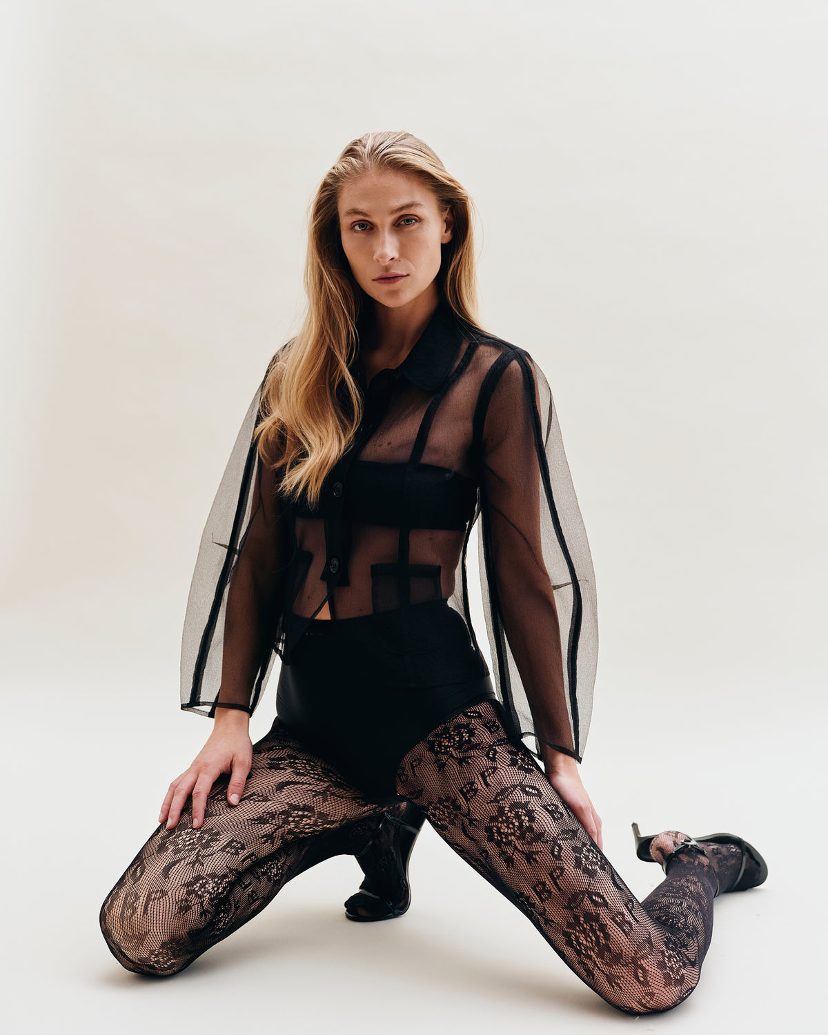Cropped campaign portrait of a seated model in a sheer black striped top over a black bustier and patterned lace tights.