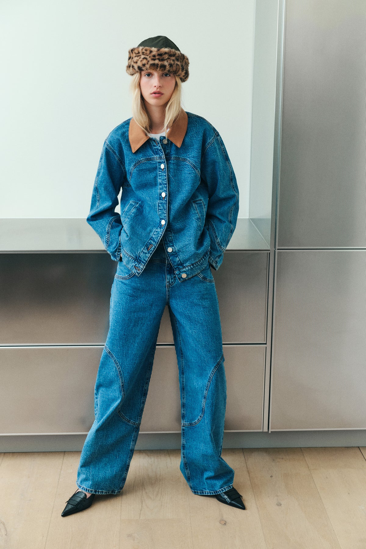 Model wearing a blue denim jacket with a tan collar, matching wide-leg jeans, and a dark hat with a leopard-print brim.