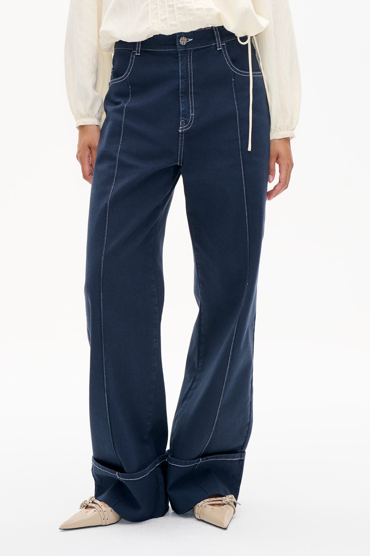 Front detail of dark indigo jeans with prominent white stitching, a long front panel, and turned-up cuffs, styled with a sheer ivory blouse.