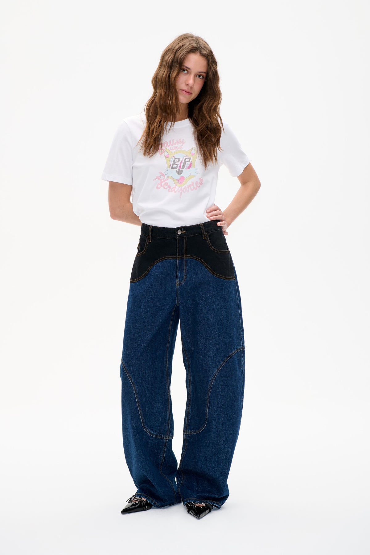 Model wearing a white short-sleeved T-shirt with a pastel cat graphic and lettering, paired with wide-leg blue jeans with black contrast panels.