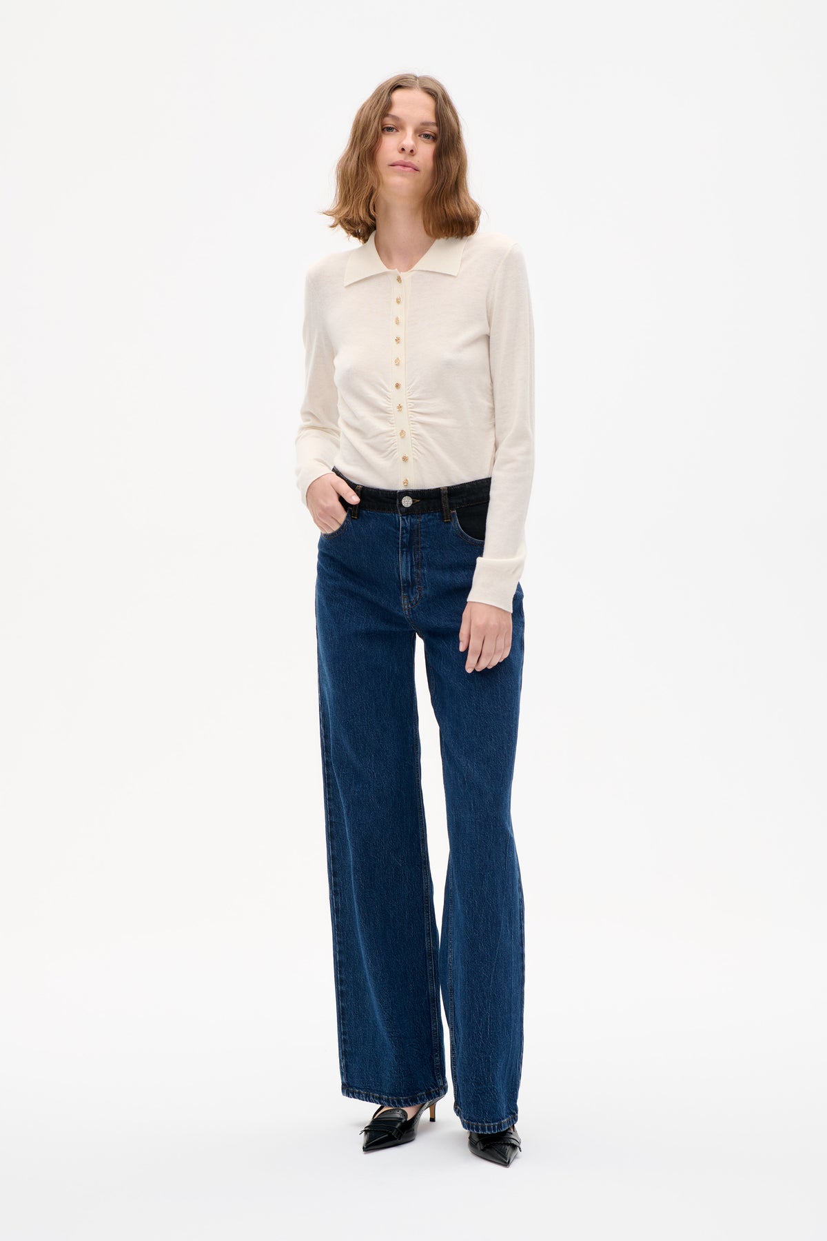 Model wearing an ivory long-sleeve cardigan with a pointed collar and decorative gold buttons, paired with dark blue wide-leg jeans.