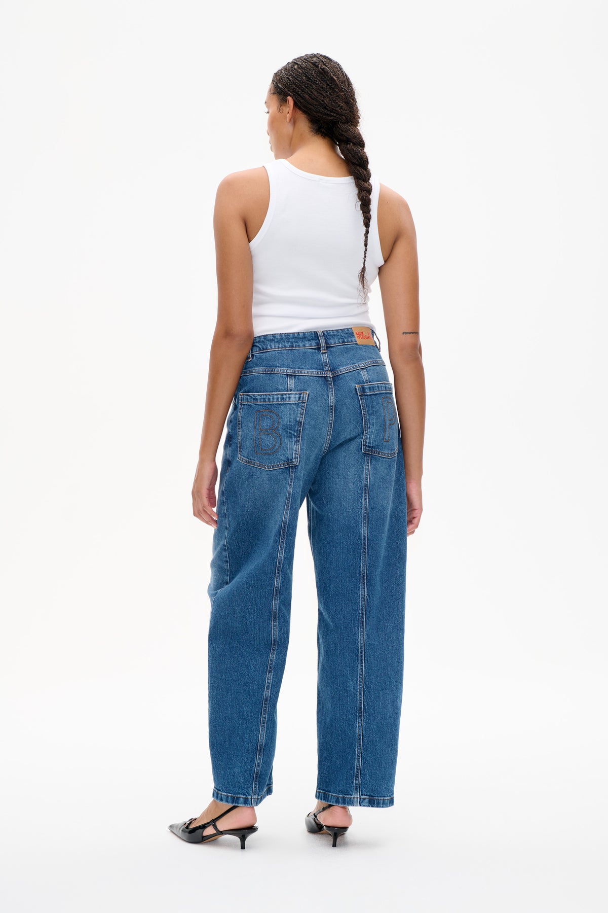 Achteraanzicht van een blauwe, wijd uitlopende jeans met grote opgestikte zakken met letters, gestyled met een witte, geribde tanktop.