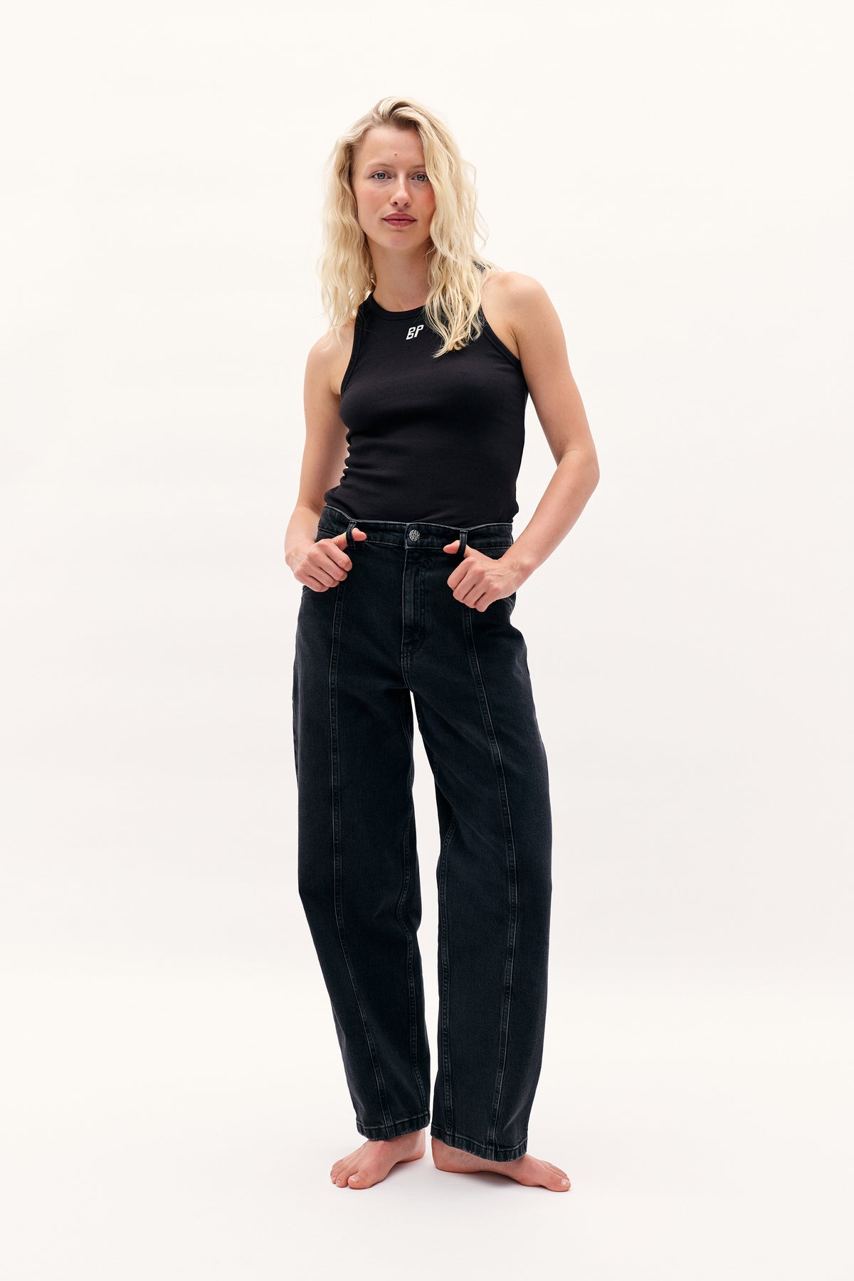 Model wearing a black sleeveless ribbed top with a small white chest logo and dark wide-leg jeans, shown from the front.