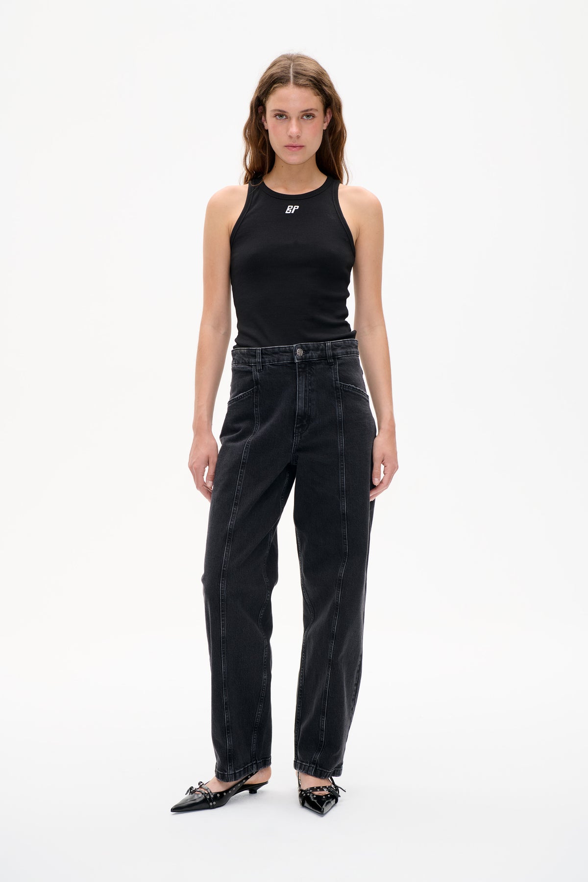 Vooraanzicht van een model in een zwarte, geribde mouwloze top met een klein wit borstlogo, gecombineerd met antracietgewassen jeans.