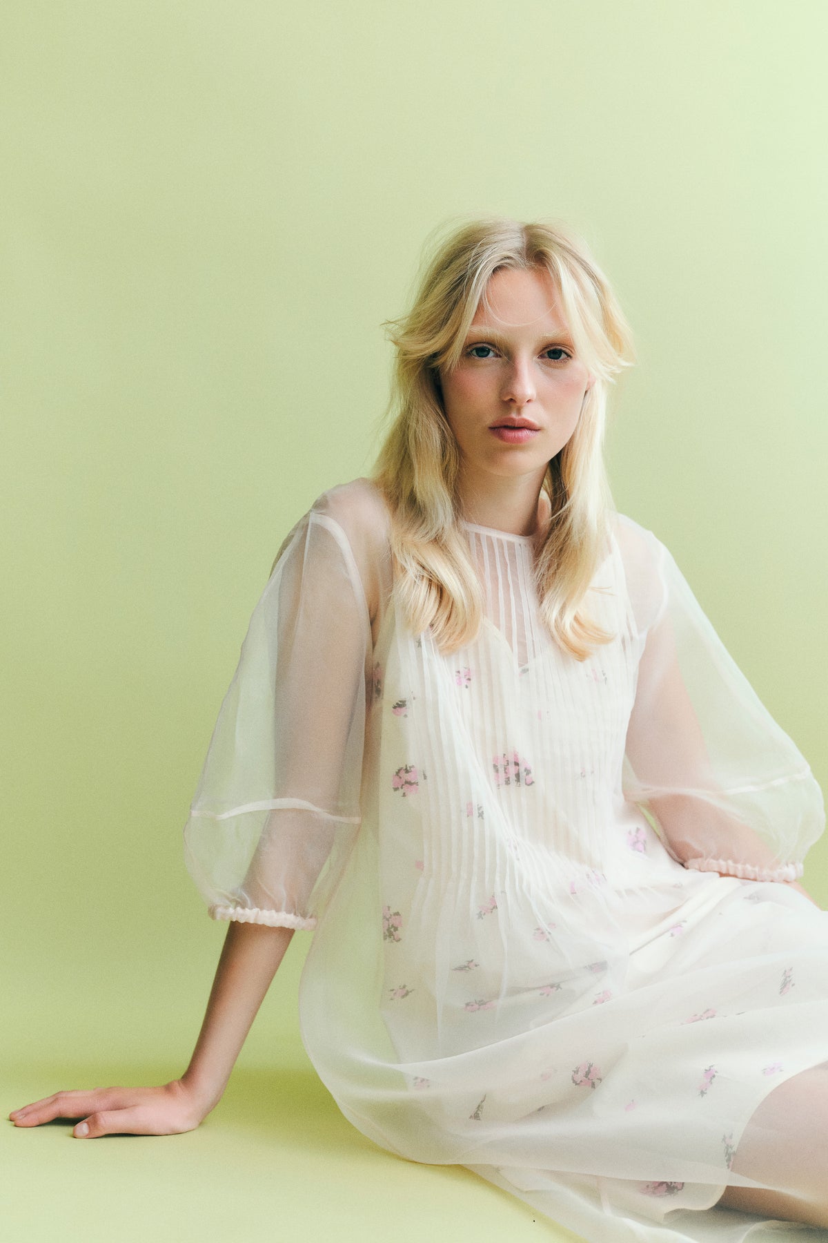 Model seated against a pale green backdrop in a white dress with small pink floral motifs and a sheer, voluminous sleeve overlay.