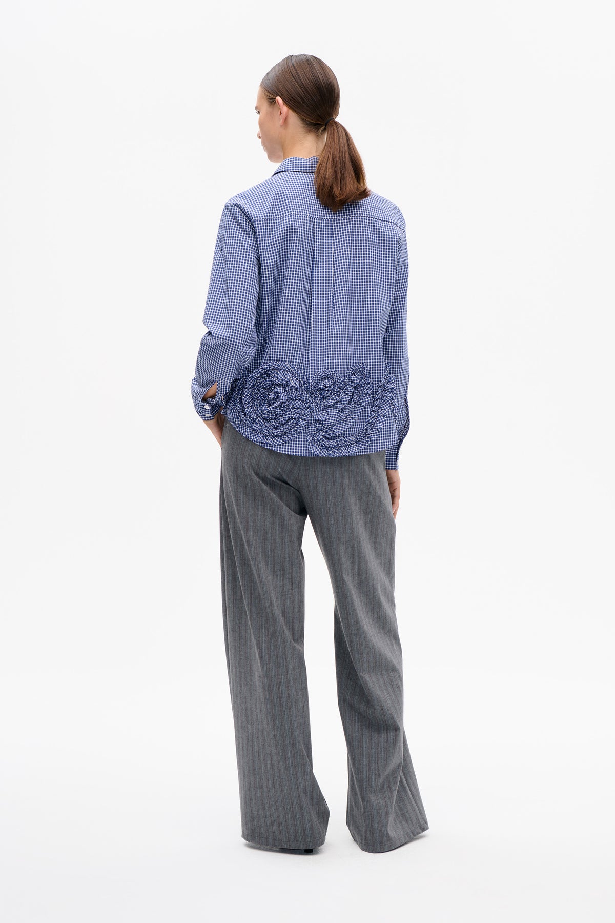 Back view of a blue-and-white gingham long-sleeved shirt with a ruffled hem, worn with grey wide-leg trousers.