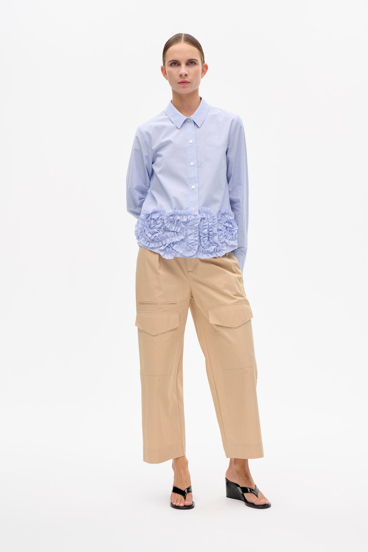 Model wearing a pale blue pinstripe button-up shirt with a chest pocket and voluminous ruffled hem, paired with beige cropped cargo trousers.