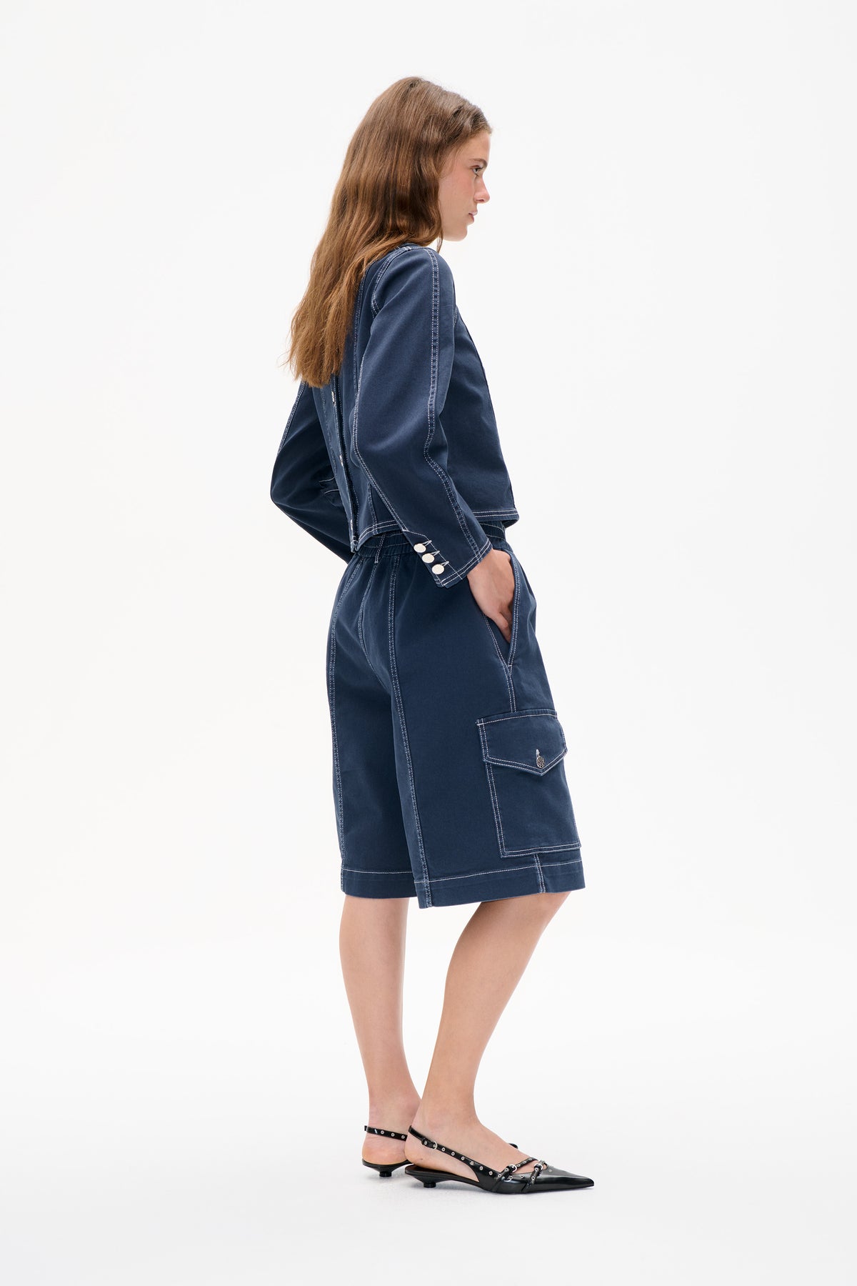 Side view of a model wearing a navy jacket and matching knee-length skirt with contrast stitching, buttoned cuffs and a large patch pocket