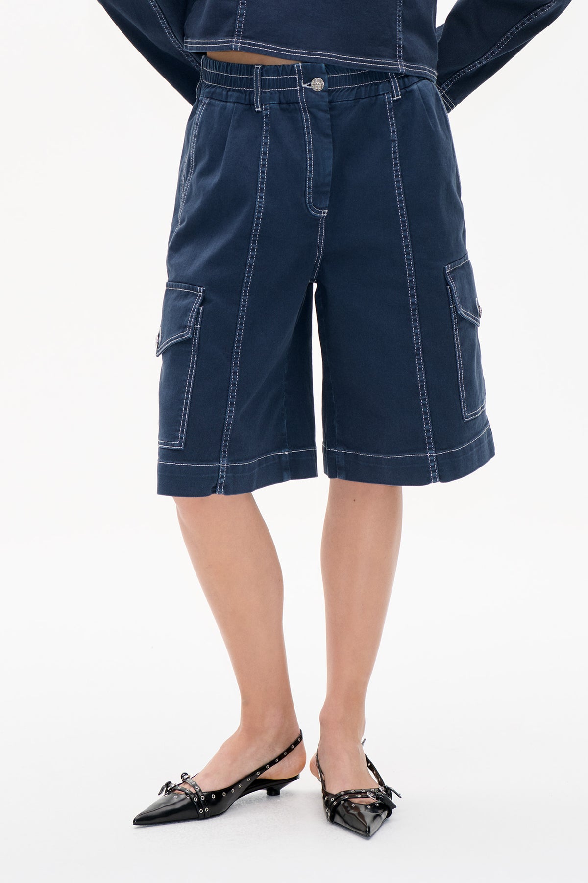 Front detail of dark indigo denim cargo shorts with an elasticated waistband, white contrast stitching and large side pockets.