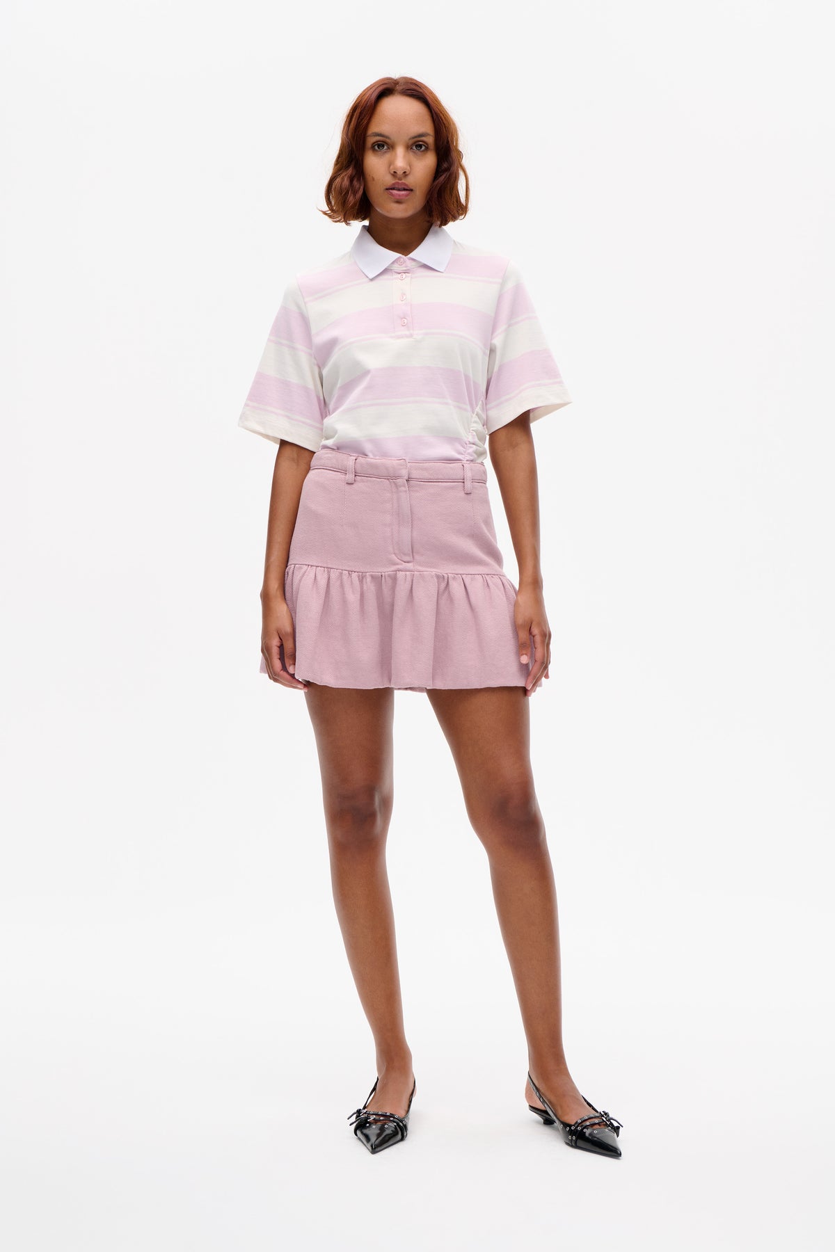 Model wearing a cream and pale pink striped short-sleeve polo with a white collar, paired with a pink ruffled mini skirt.