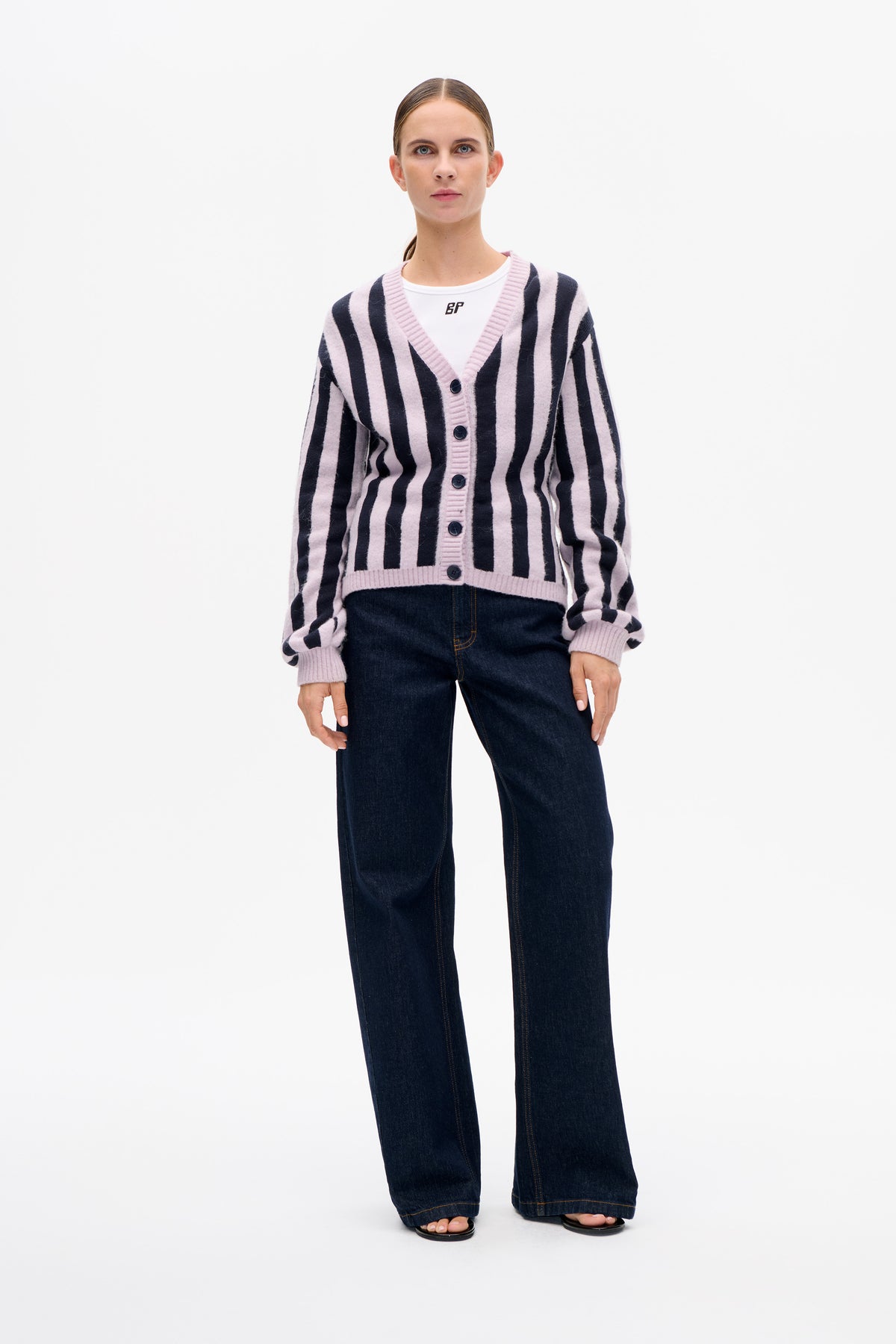 Front view of a model wearing a pale pink and navy striped button-front cardigan over a white T-shirt and dark wide-leg jeans.