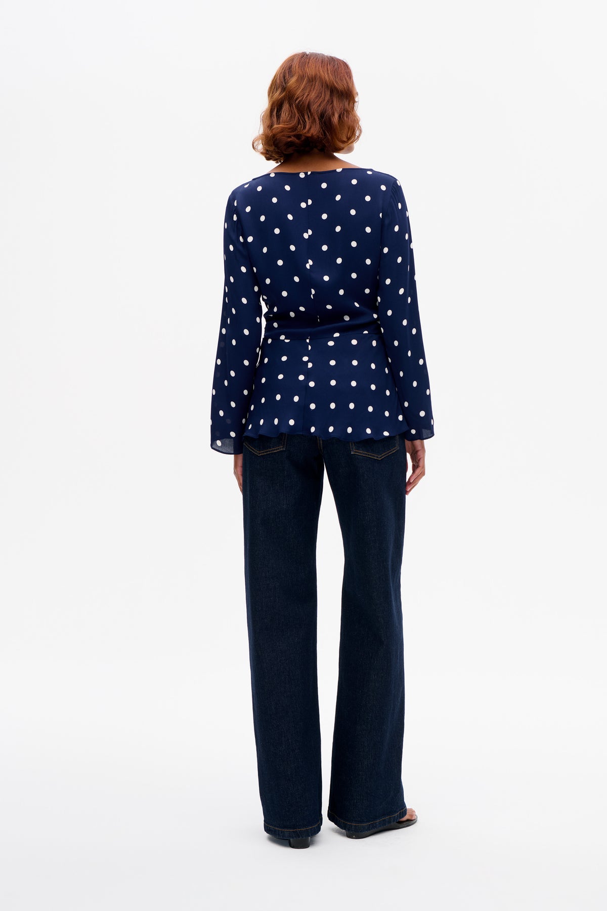 Back view of a navy long-sleeved blouse with white polka dots and a flared hem, paired with dark wide-leg jeans.