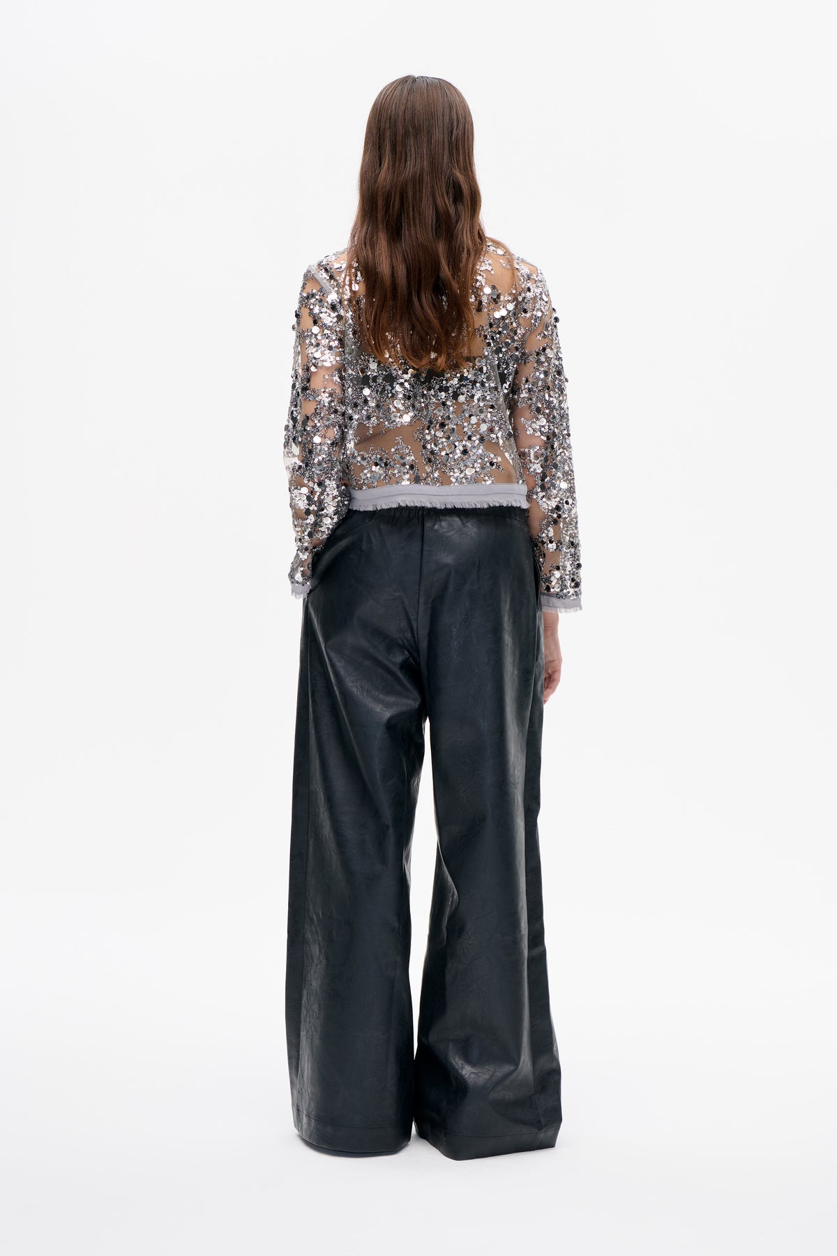 Back view of a model wearing a sheer long-sleeved top covered in silver and black sequins with black wide-leg trousers.